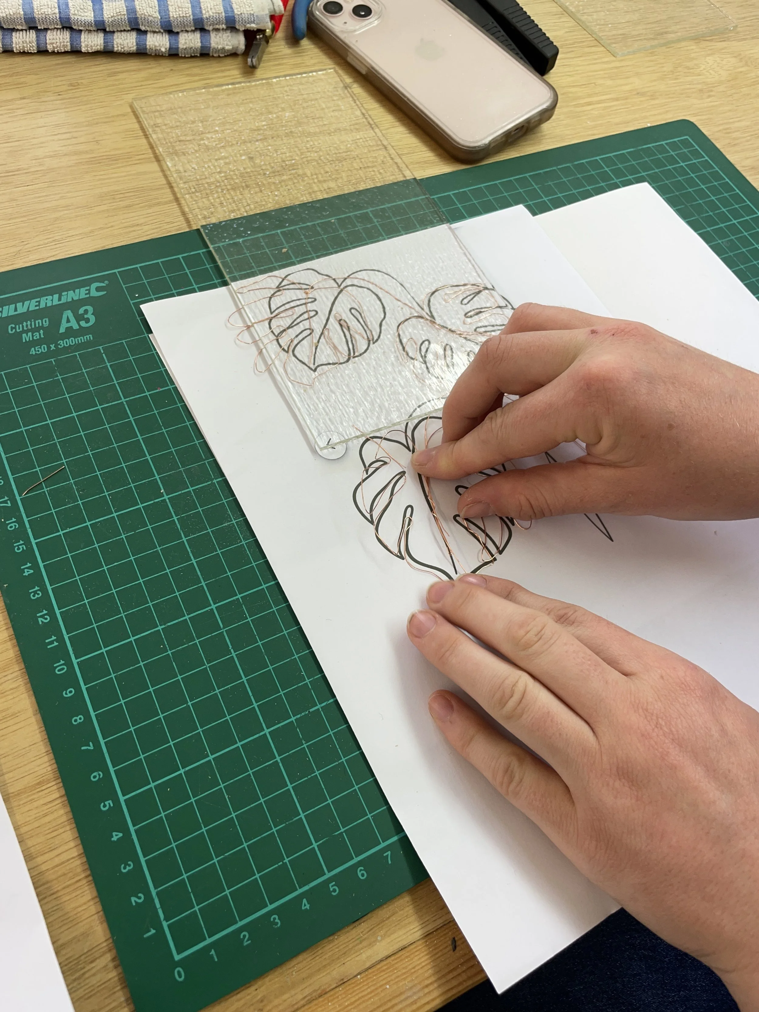 Person tracing leaves on paper placed on a cutting mat with a smartphone nearby.
