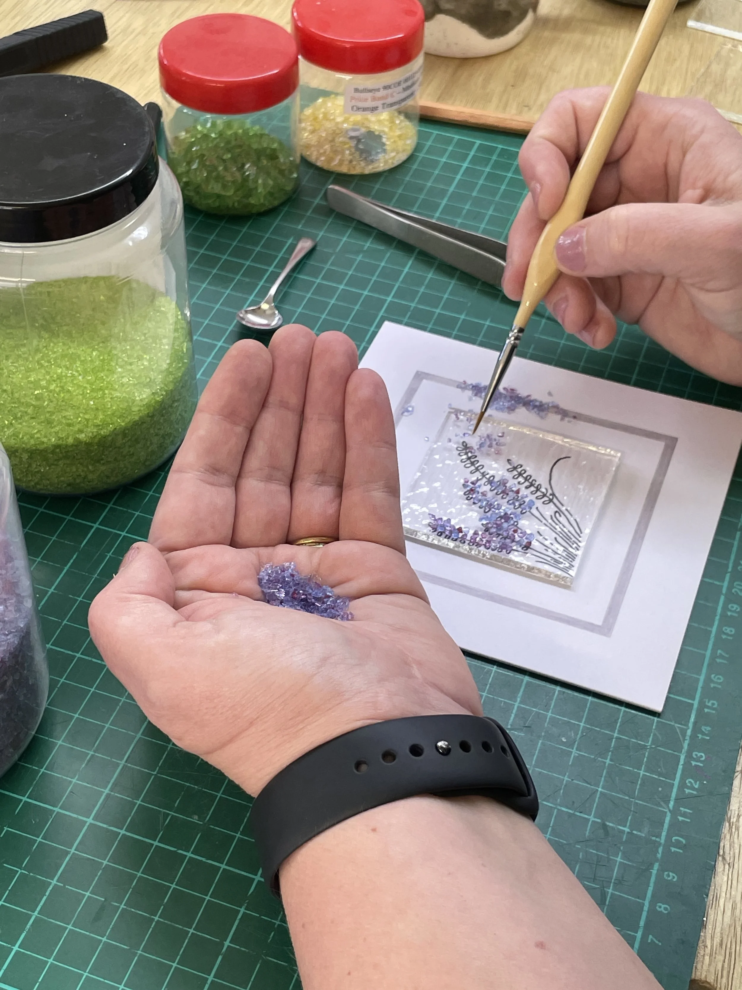 Person crafting with colorful glass fragments on a mat, using a brush to arrange pieces on a paper template. Various jars filled with glass pieces and a spoon are on the table.