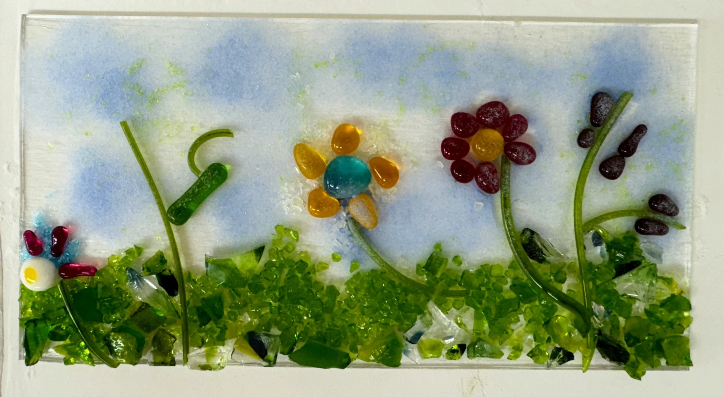 Glass artwork depicting a colorful floral scene with green leaves, a blue sky background, and vibrant flowers made from colorful glass pieces.