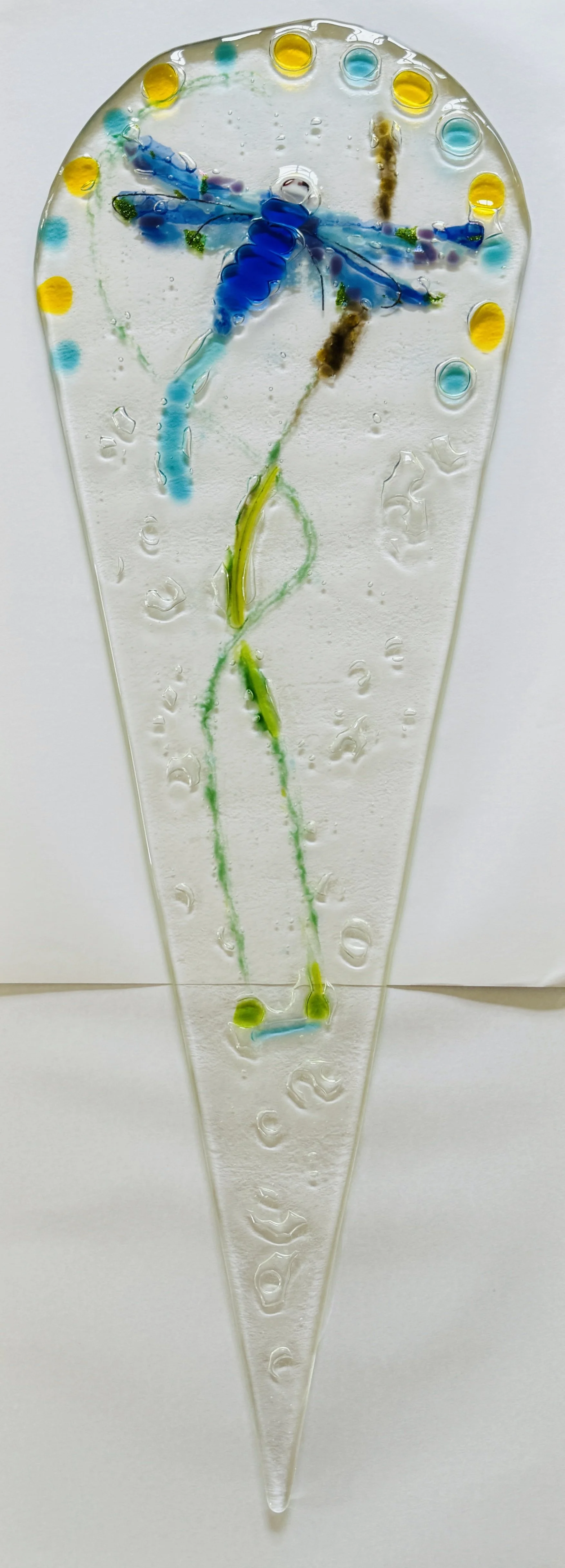 Glass art depicting a blue dragonfly with a green stem, surrounded by yellow and blue dots, on a light background.