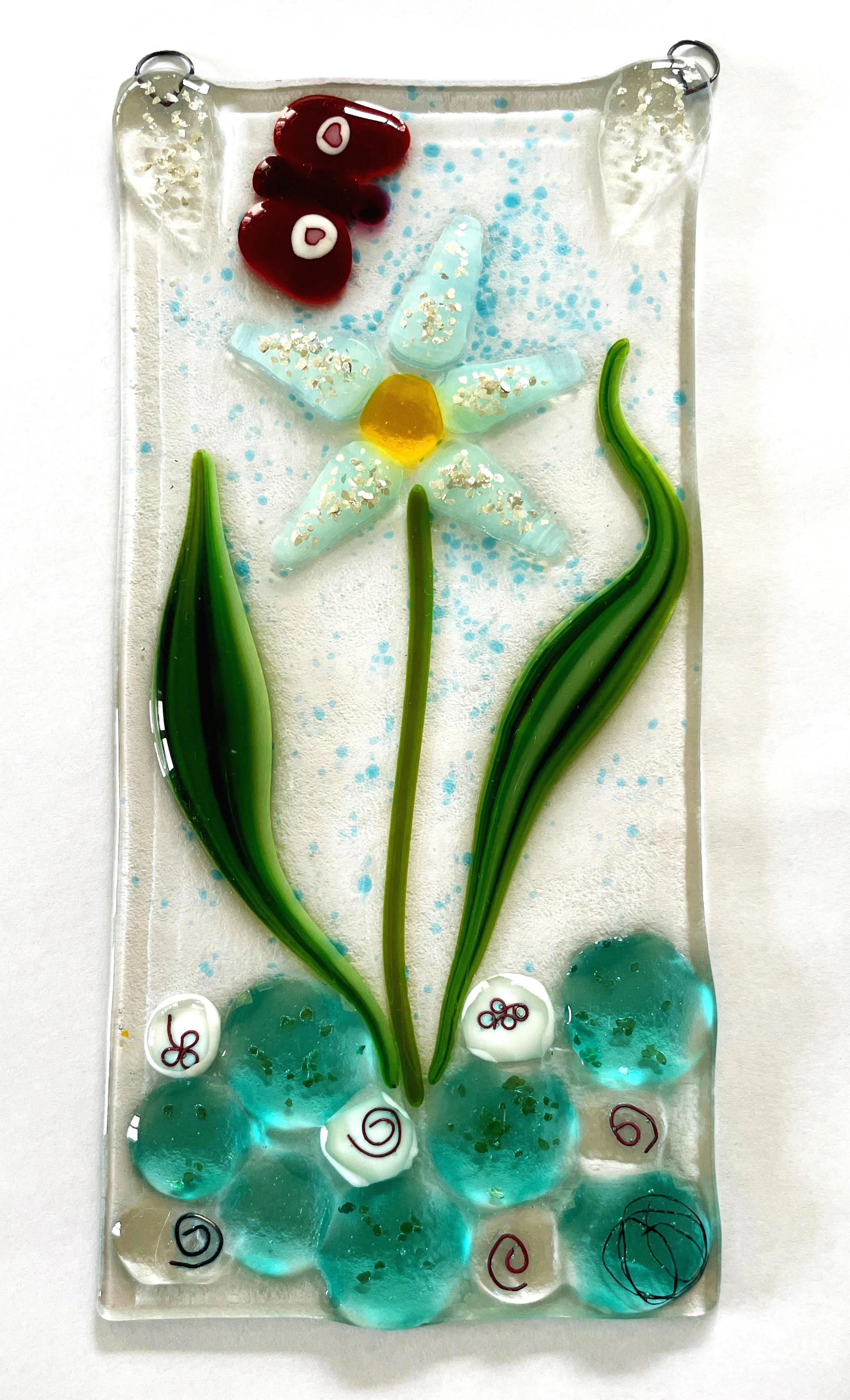 Glass art panel featuring a white flower with a yellow center, green leaves, and a red butterfly. Background includes turquoise and white decorative elements.