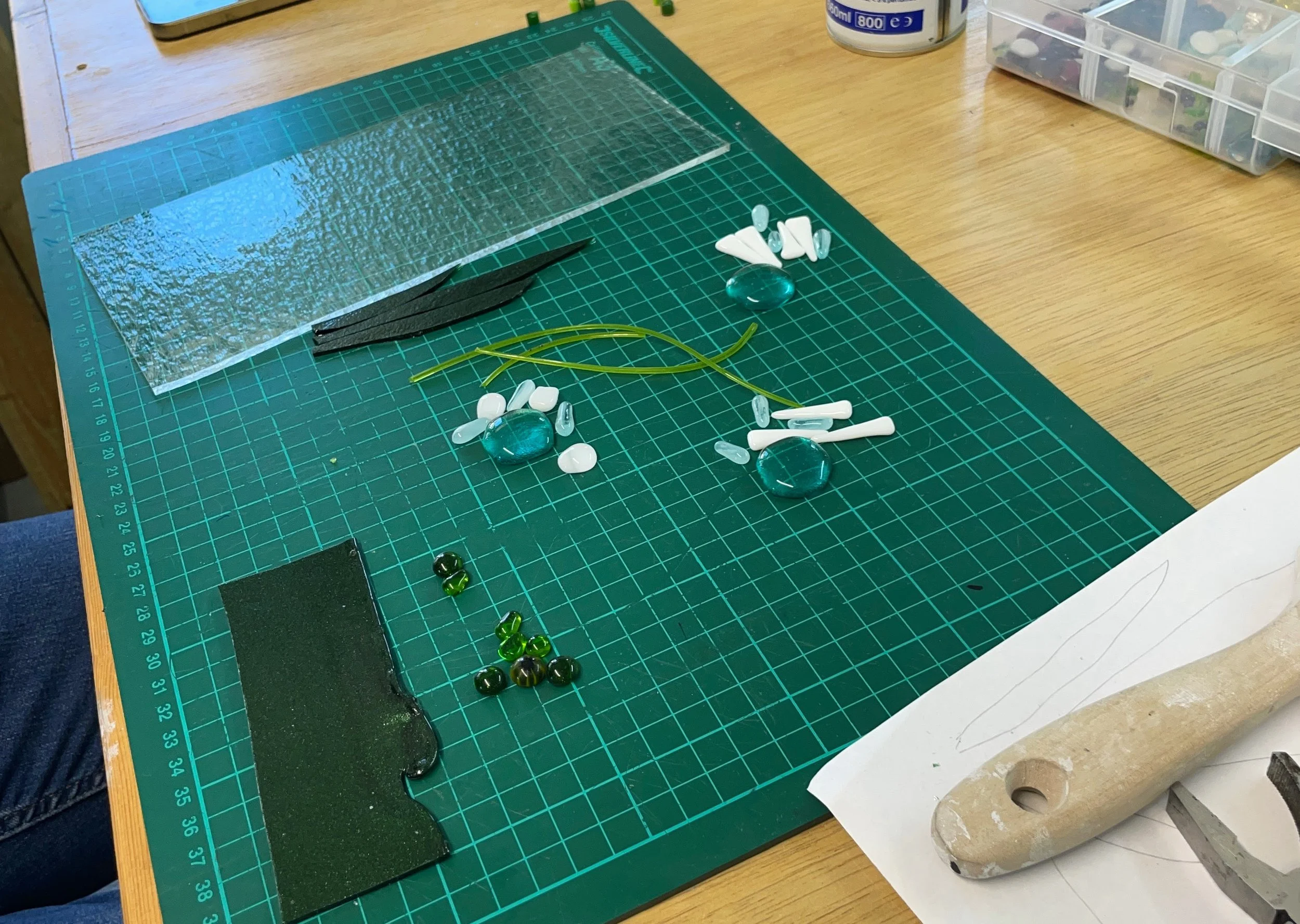 A crafting workspace with a green cutting mat, various glass pieces and beads, a wooden tool, and small plastic containers on a wooden table.