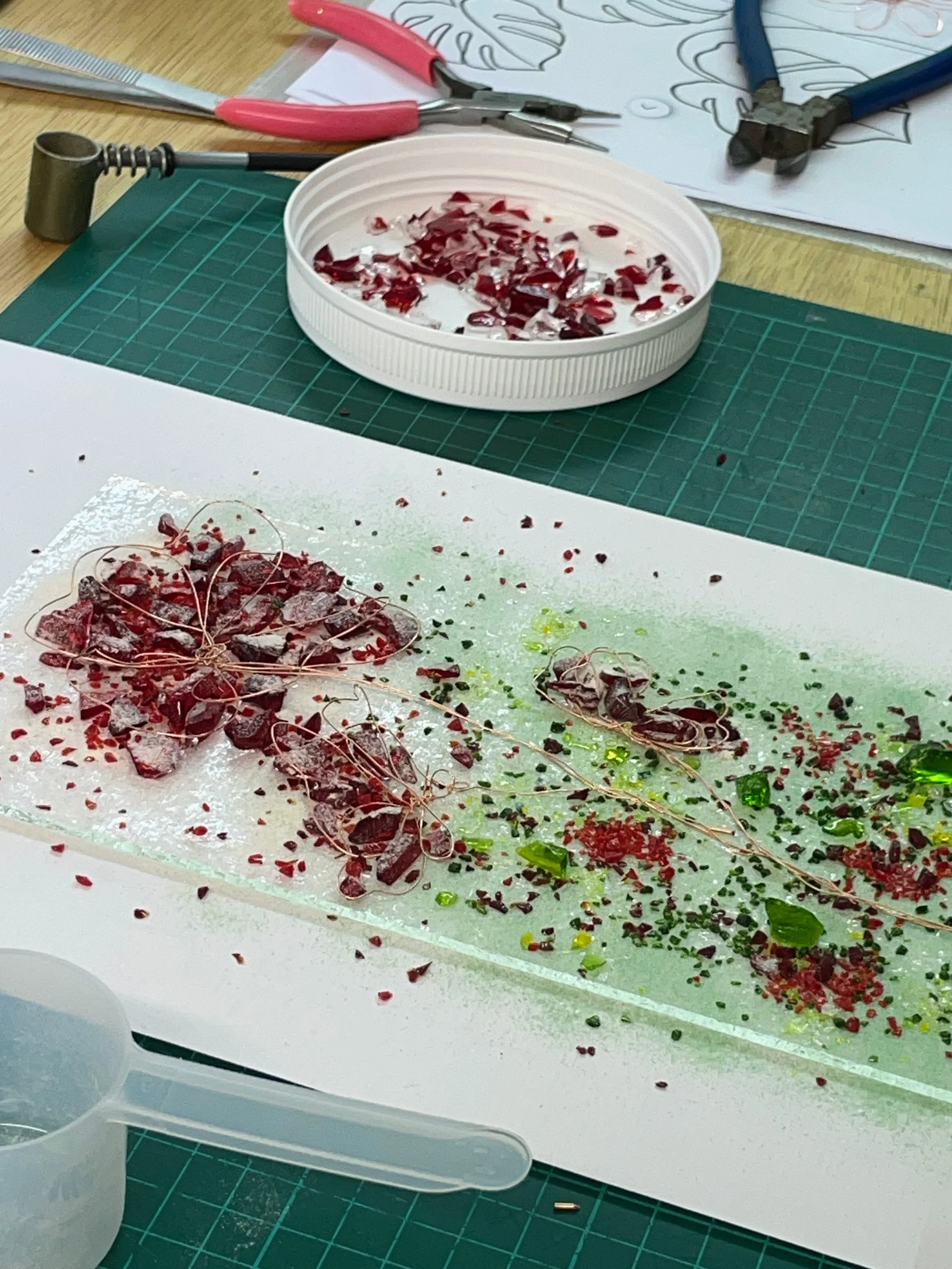 A table with red and green glass fragments, tools like pliers and a cutting board. There's a white container lid holding red glass pieces.