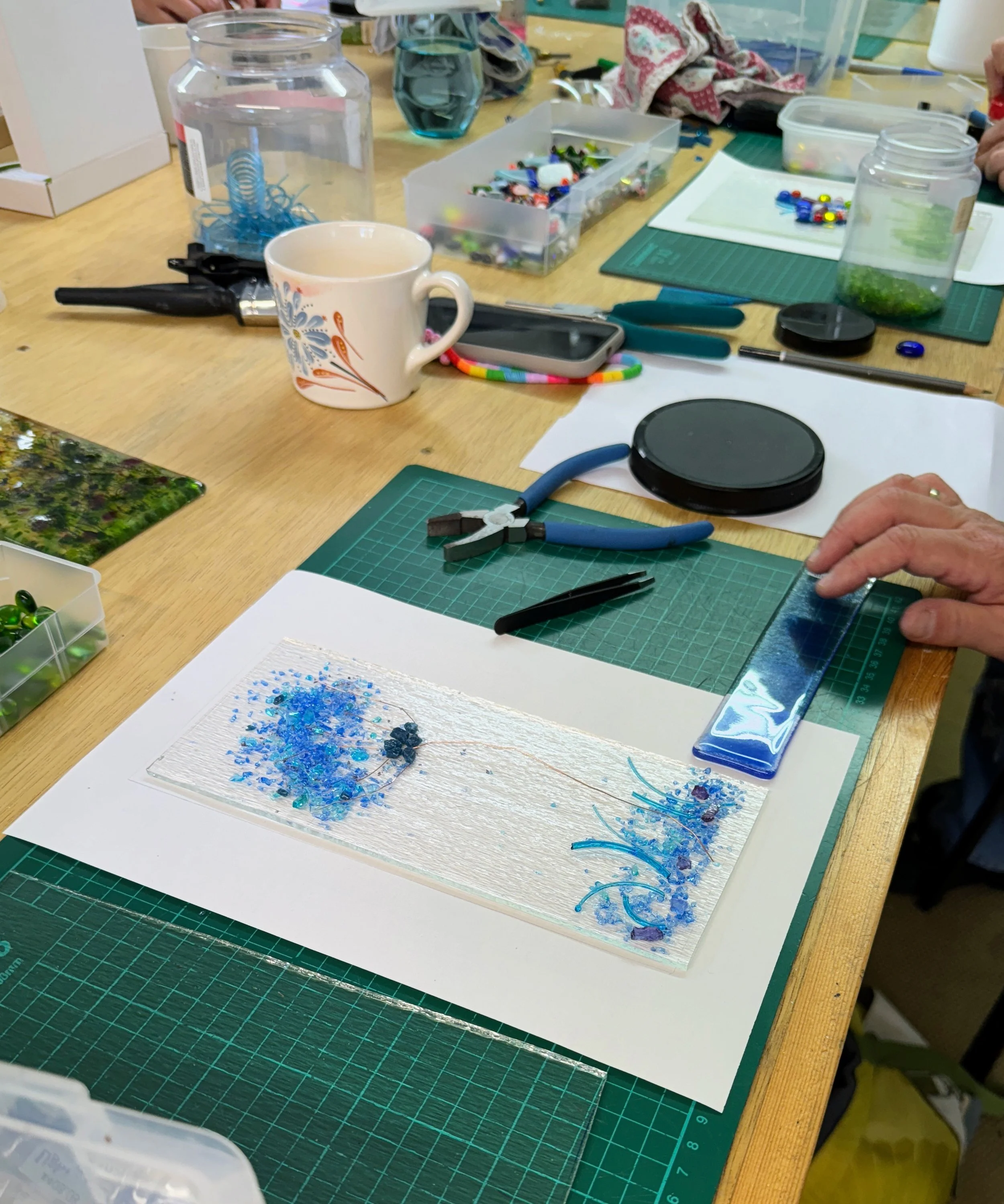 Arts and crafts workspace with tools, a piece of glass art in progress, colorful beads, and a floral mug on a table.