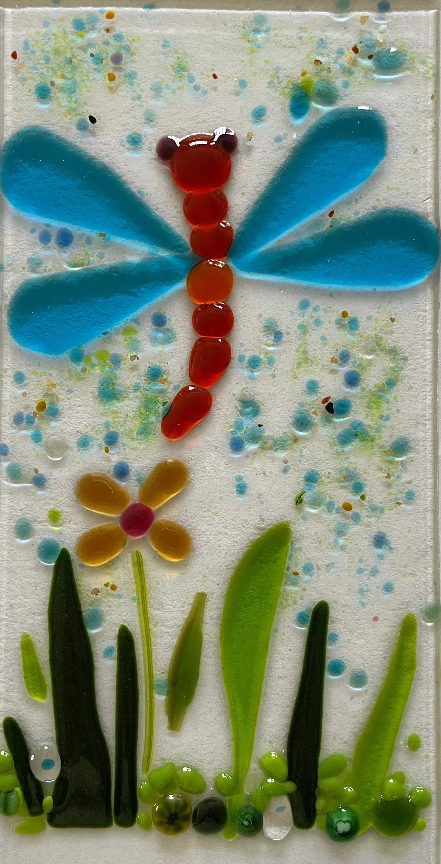 Glass artwork featuring a dragonfly with blue wings and a red body, a yellow flower with a pink center, and green grass on a speckled background.