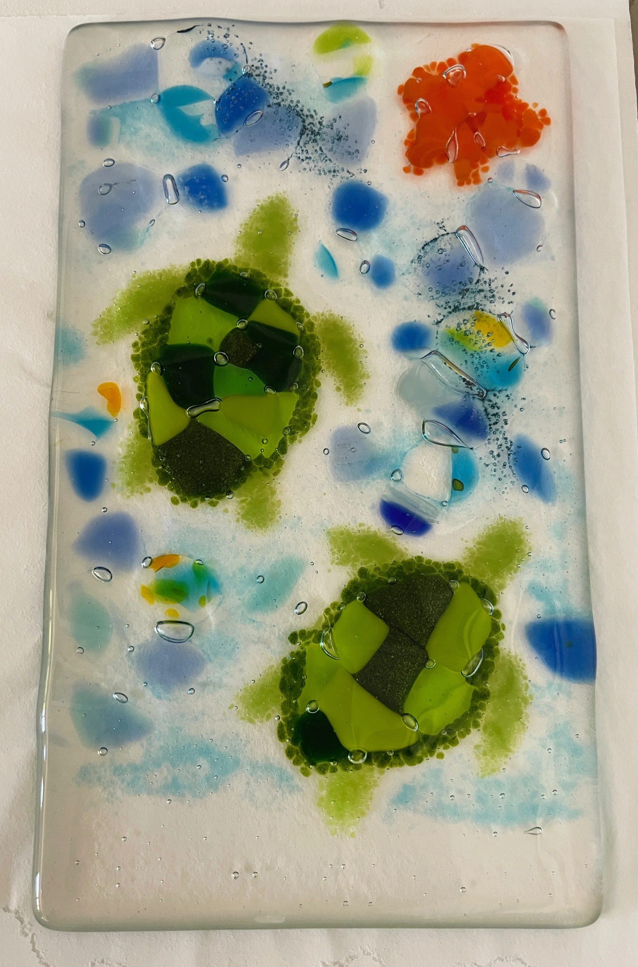 Glass artwork featuring green sea turtles swimming among colorful abstract shapes in blue, orange, and white.