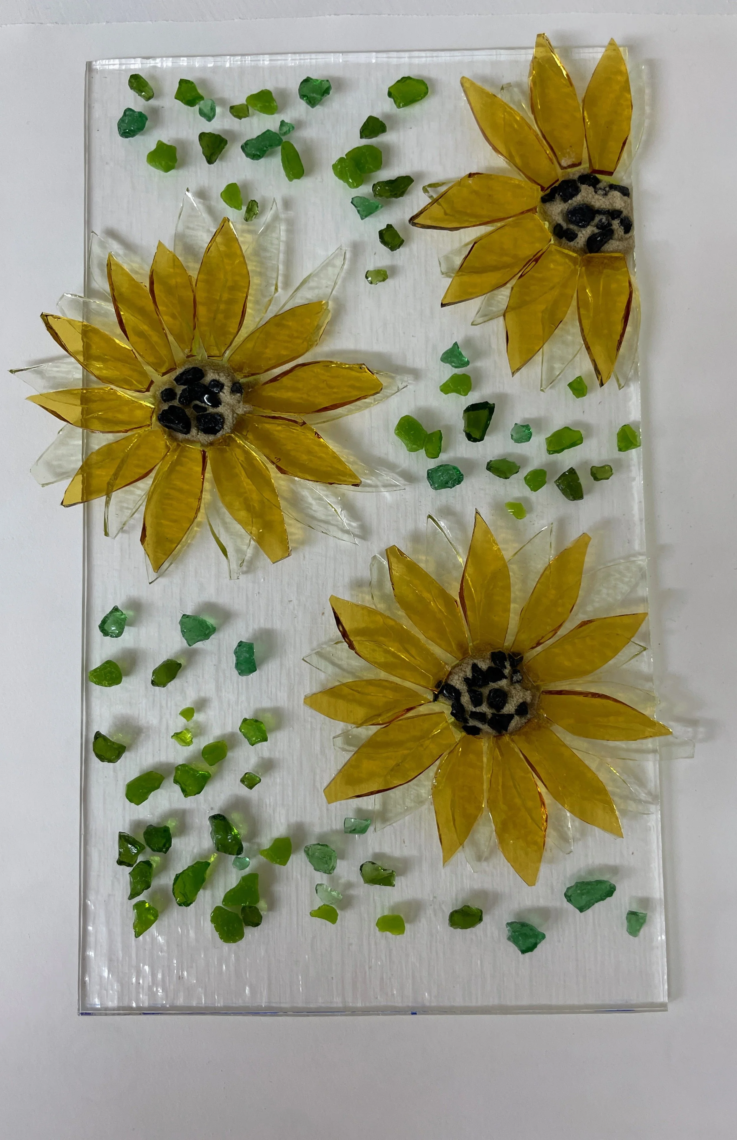 Glass artwork with three yellow sunflowers and green pieces on a clear background.