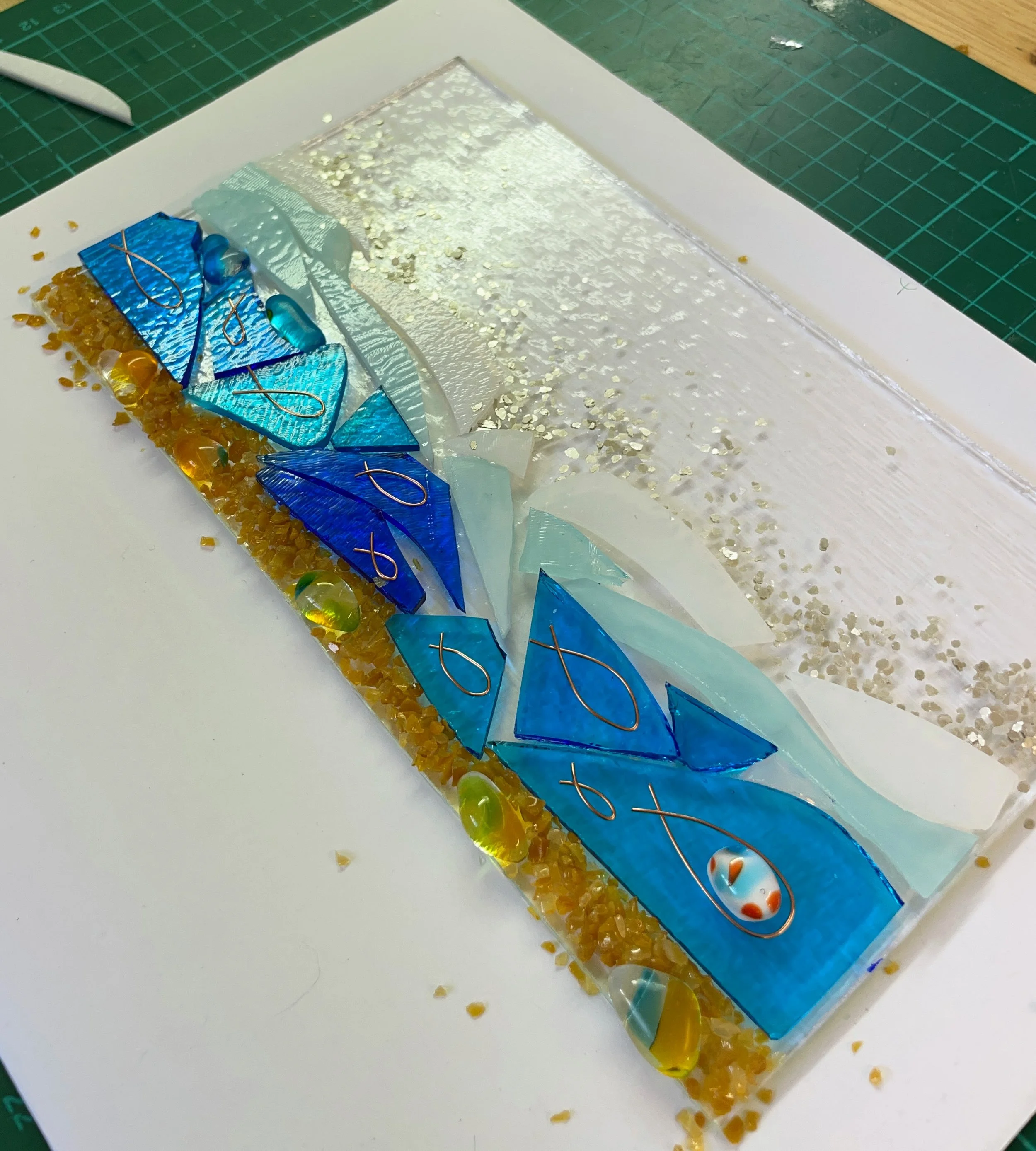 Colorful mosaic art with blue and turquoise triangular pieces, resembling fish, combined with gold accents and transparent pebbles on a textured background.