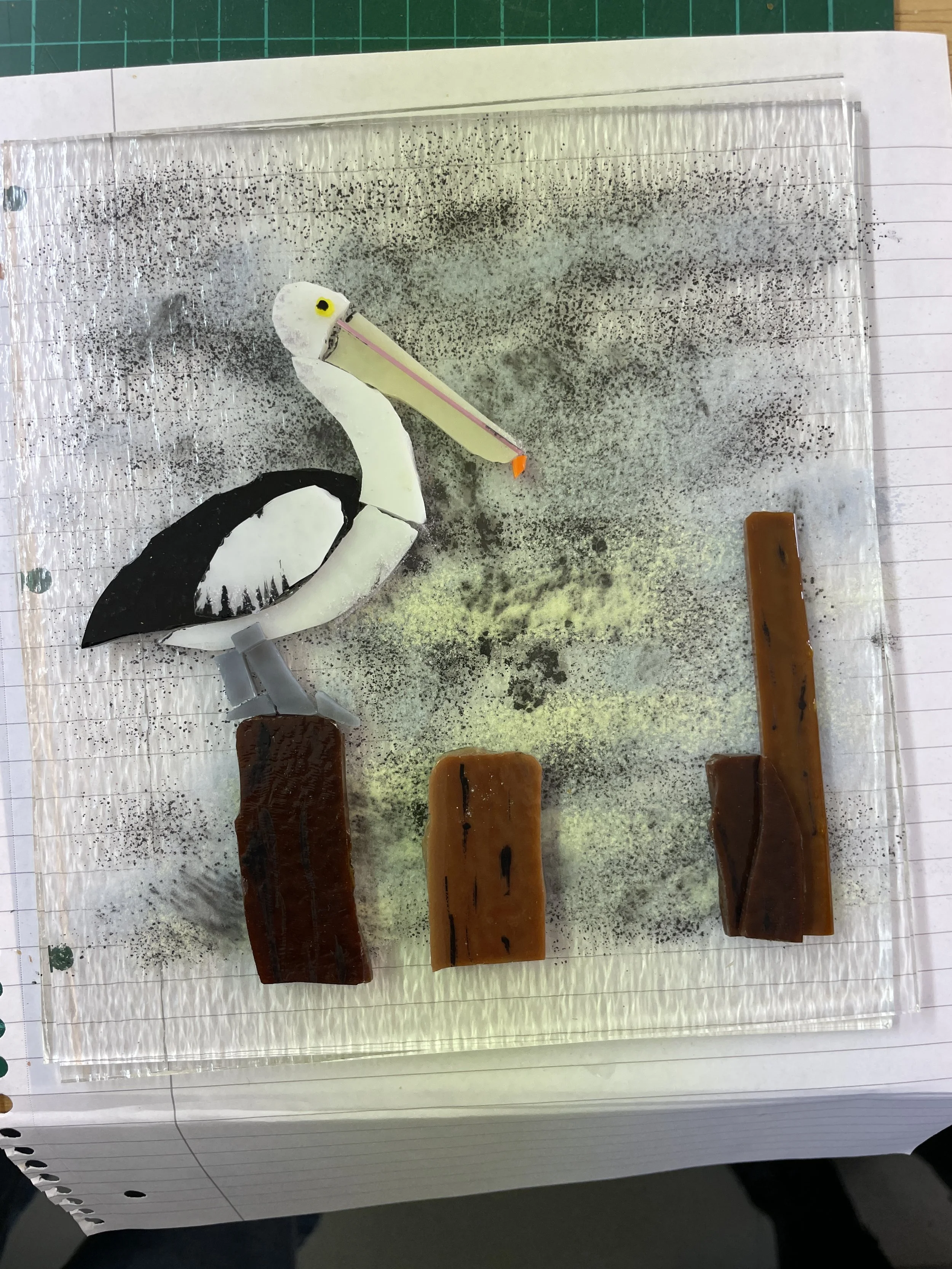 Glass art piece featuring a pelican with a long beak perched on a brown post, surrounded by abstract textures and colors on a notepad background.