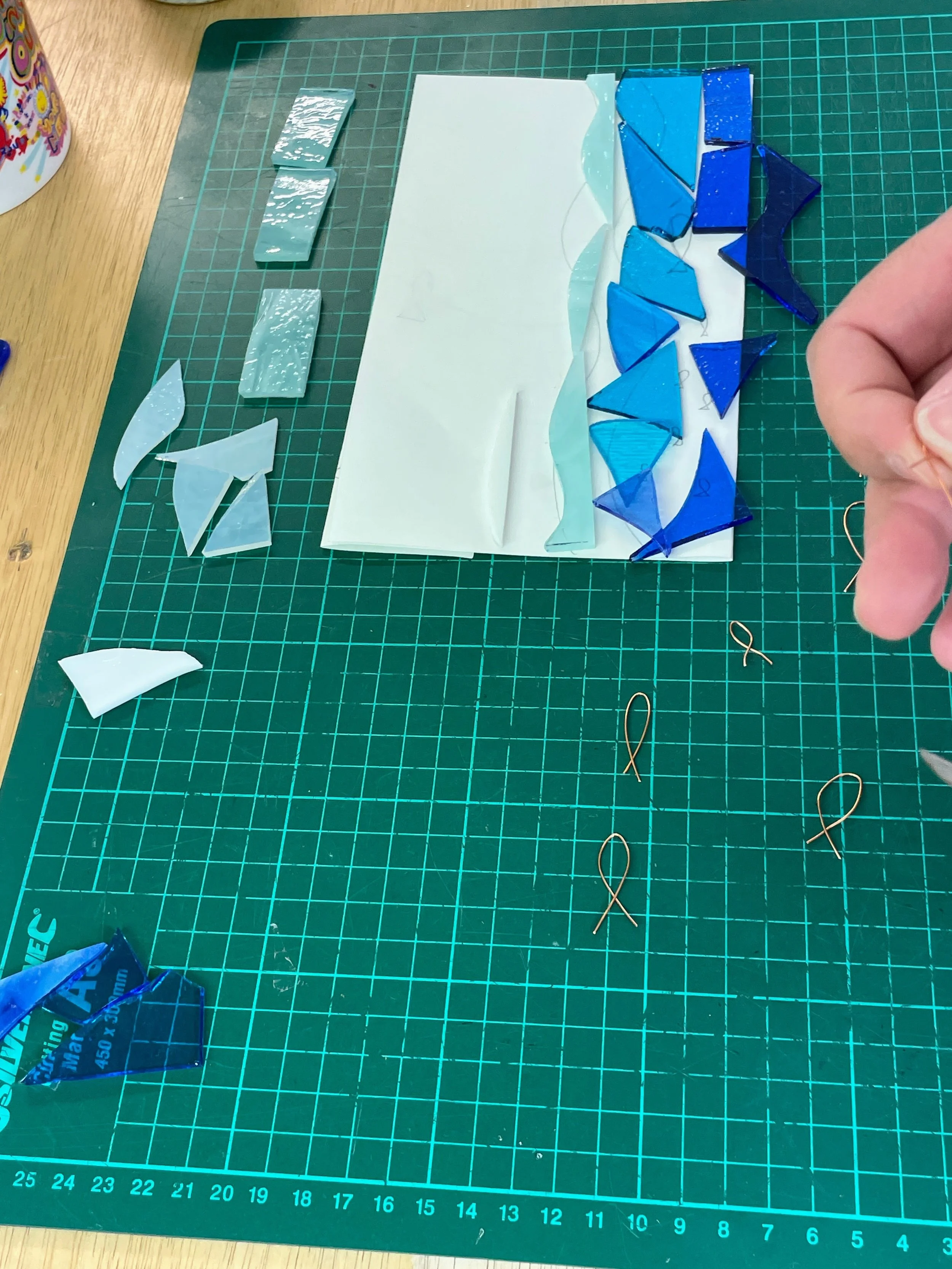 Crafting workspace with blue and white stained glass pieces on a green cutting mat, alongside copper wire loops and tools.