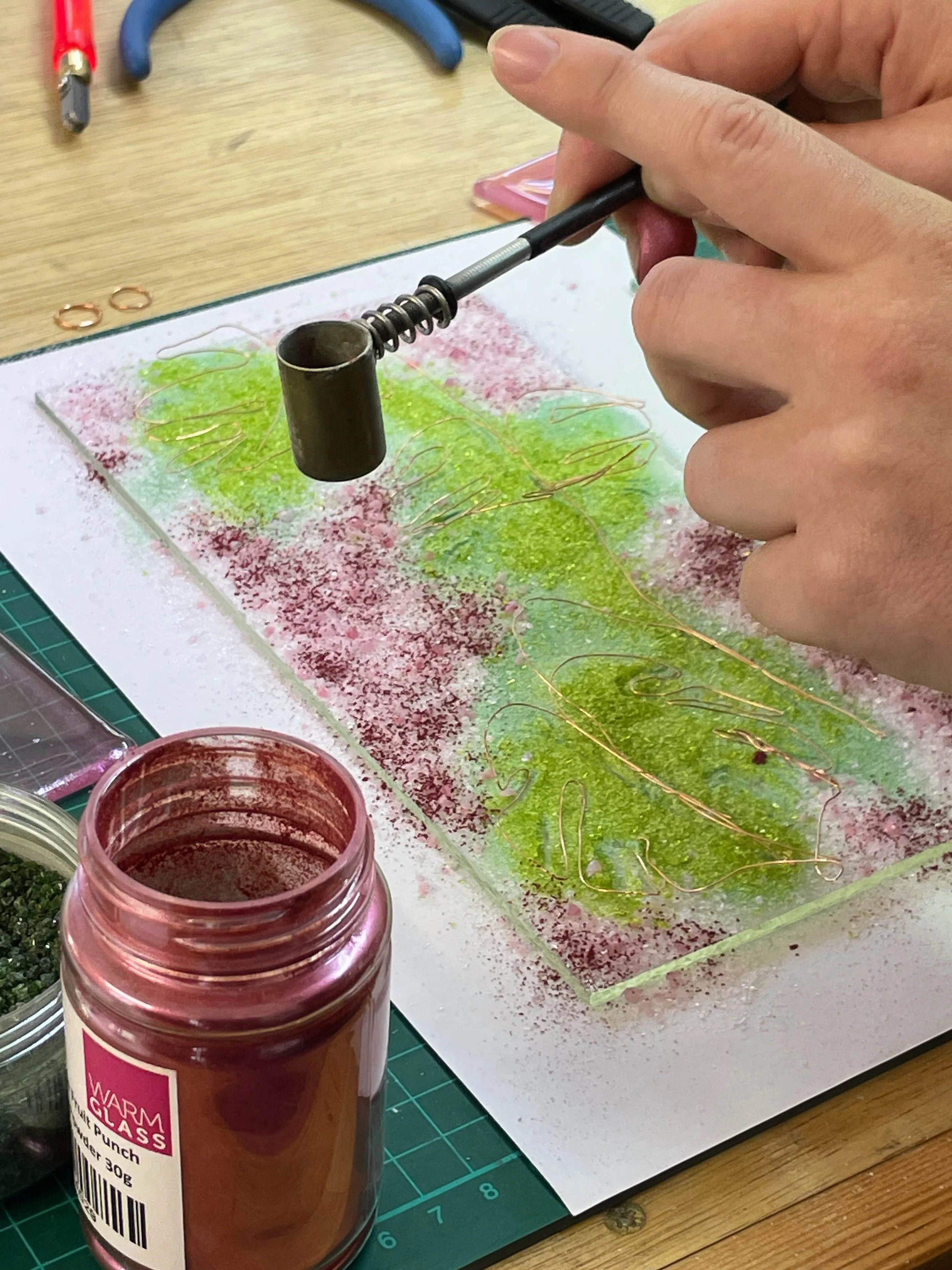 Person using a tool to work with colored glass pieces on a work surface. There is a jar of red powder and green and pink glass fragments. Wire and tools are visible nearby, indicating a craft or art project in progress.