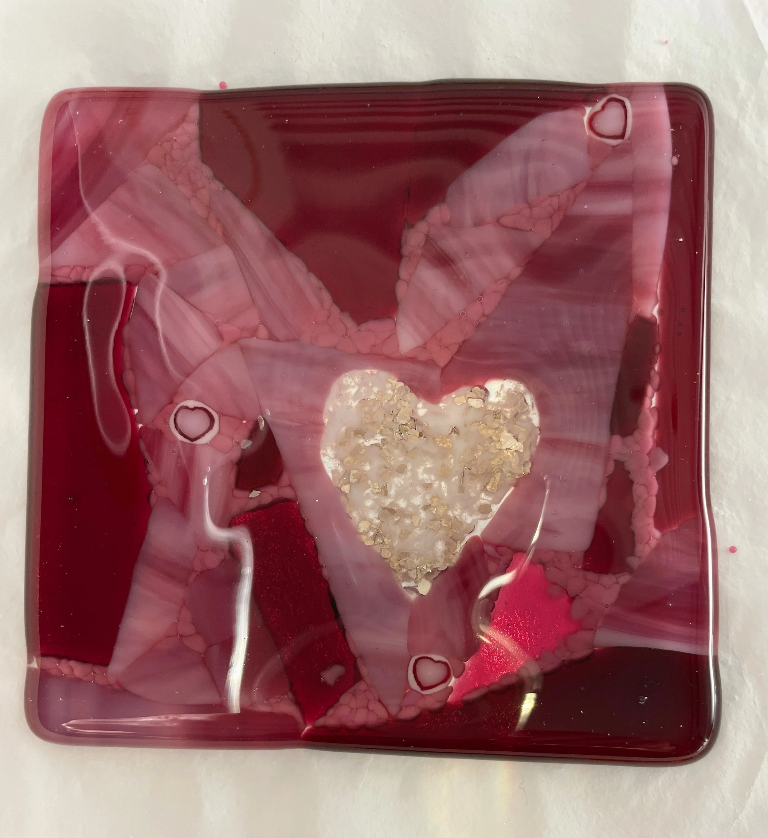Square glass art piece featuring a red base with swirling pink and white patterns, centered by a heart-shaped design with gold flecks.