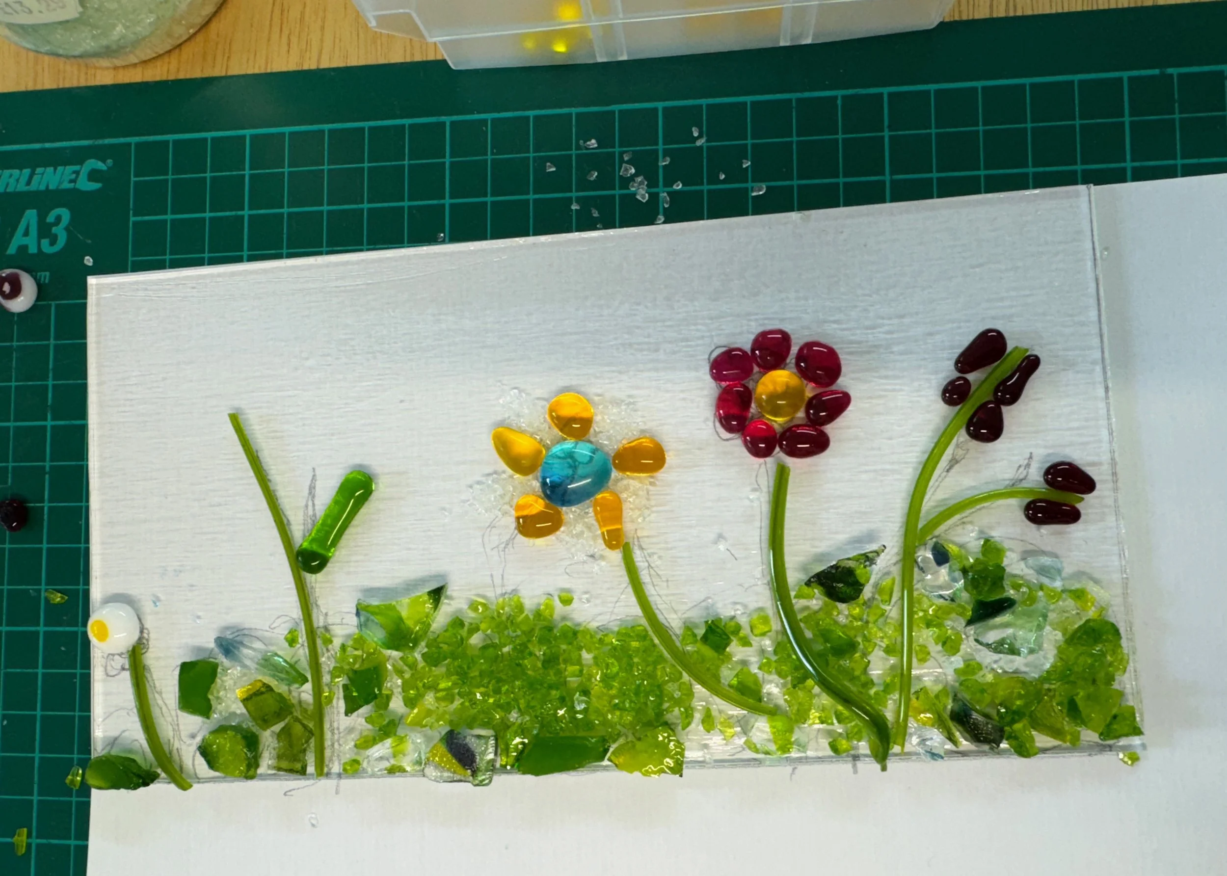Fused glass artwork depicting flowers with colorful glass pieces on a grid mat.
