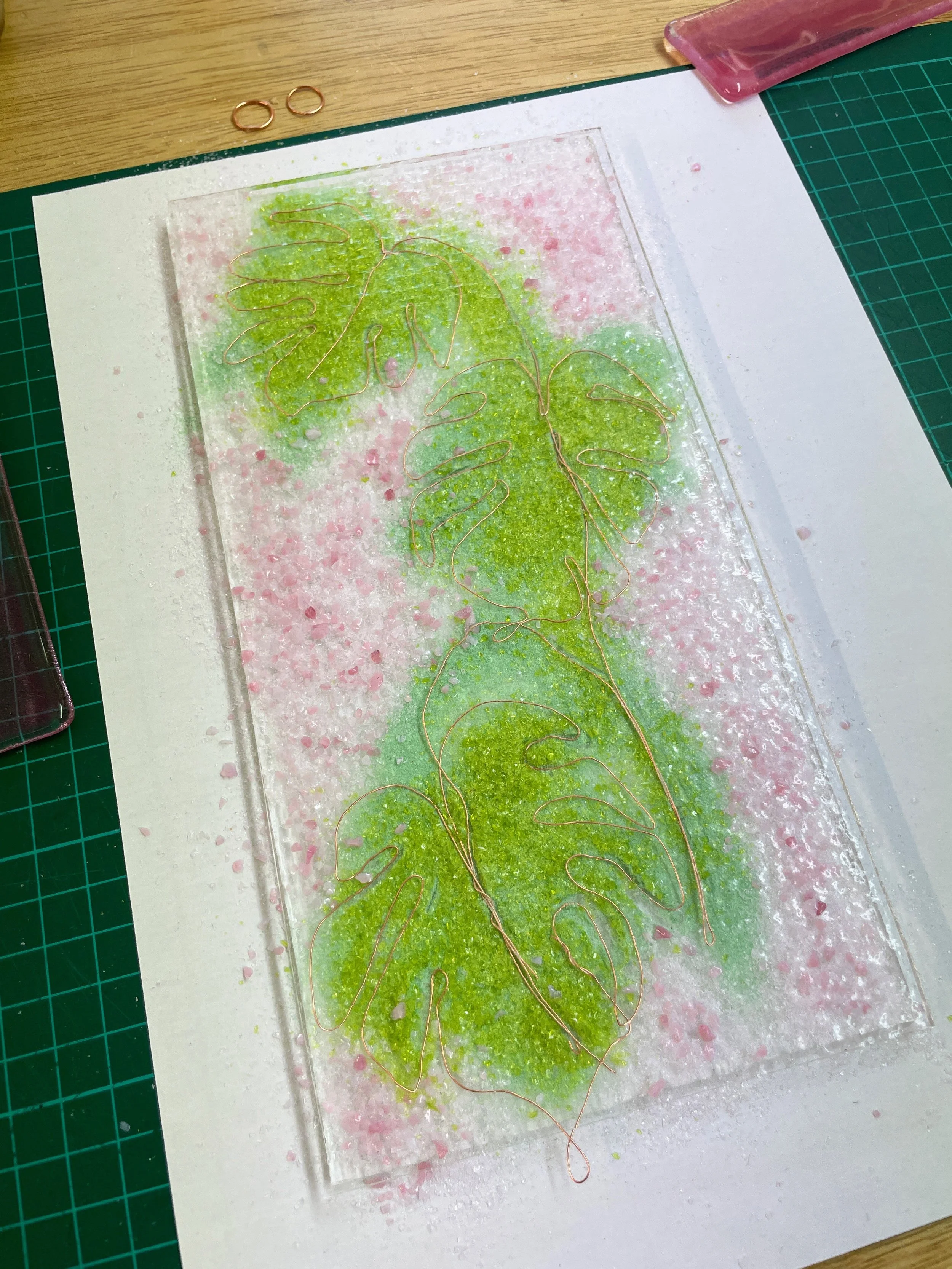Glass artwork with green and pink textured background, featuring wire outlines of tropical leaves on a table with a cutting mat.