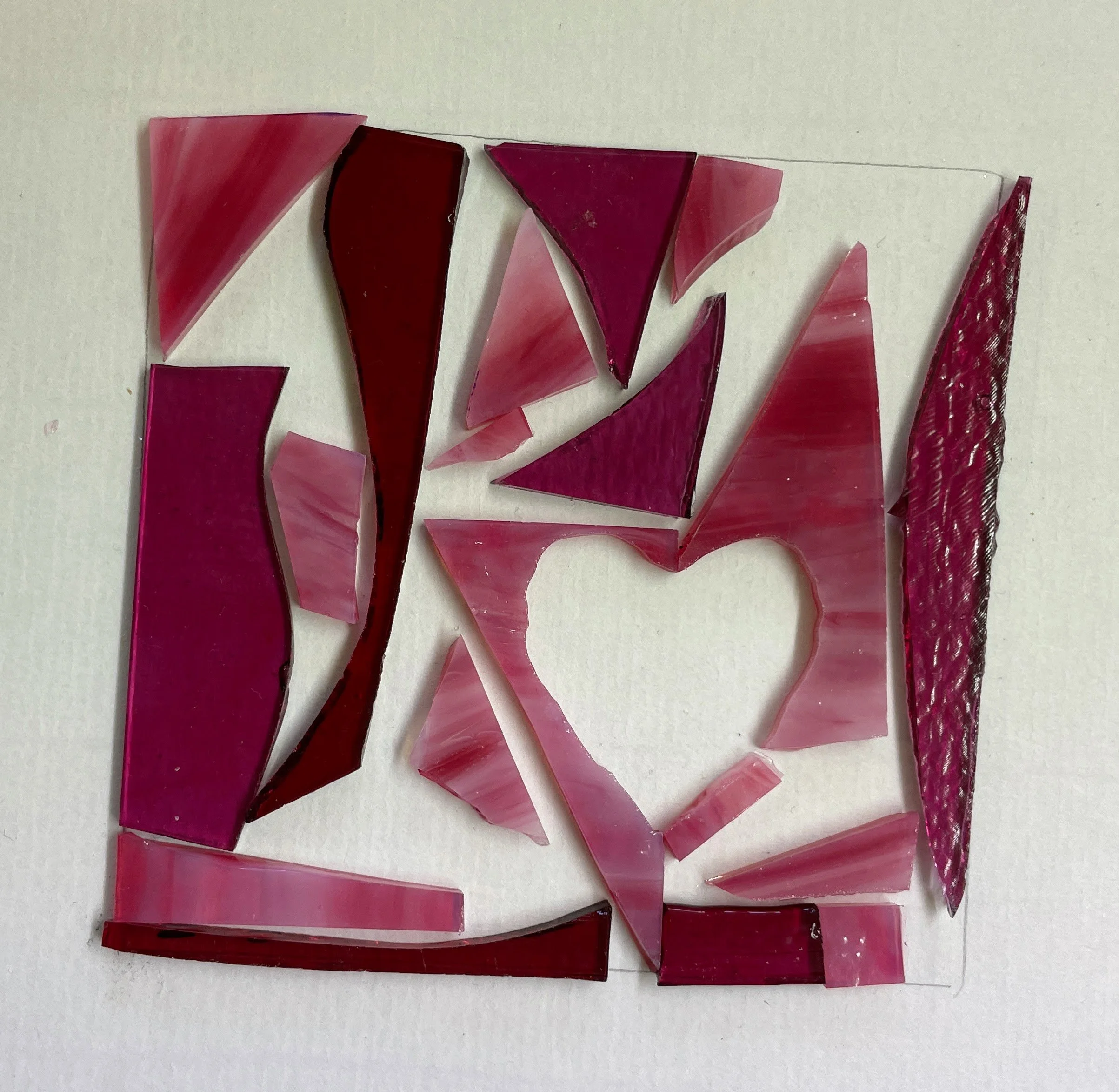 Colorful stained glass pieces arranged on a white background, with varying shades of red and pink, forming geometric shapes and a heart cutout in the center.