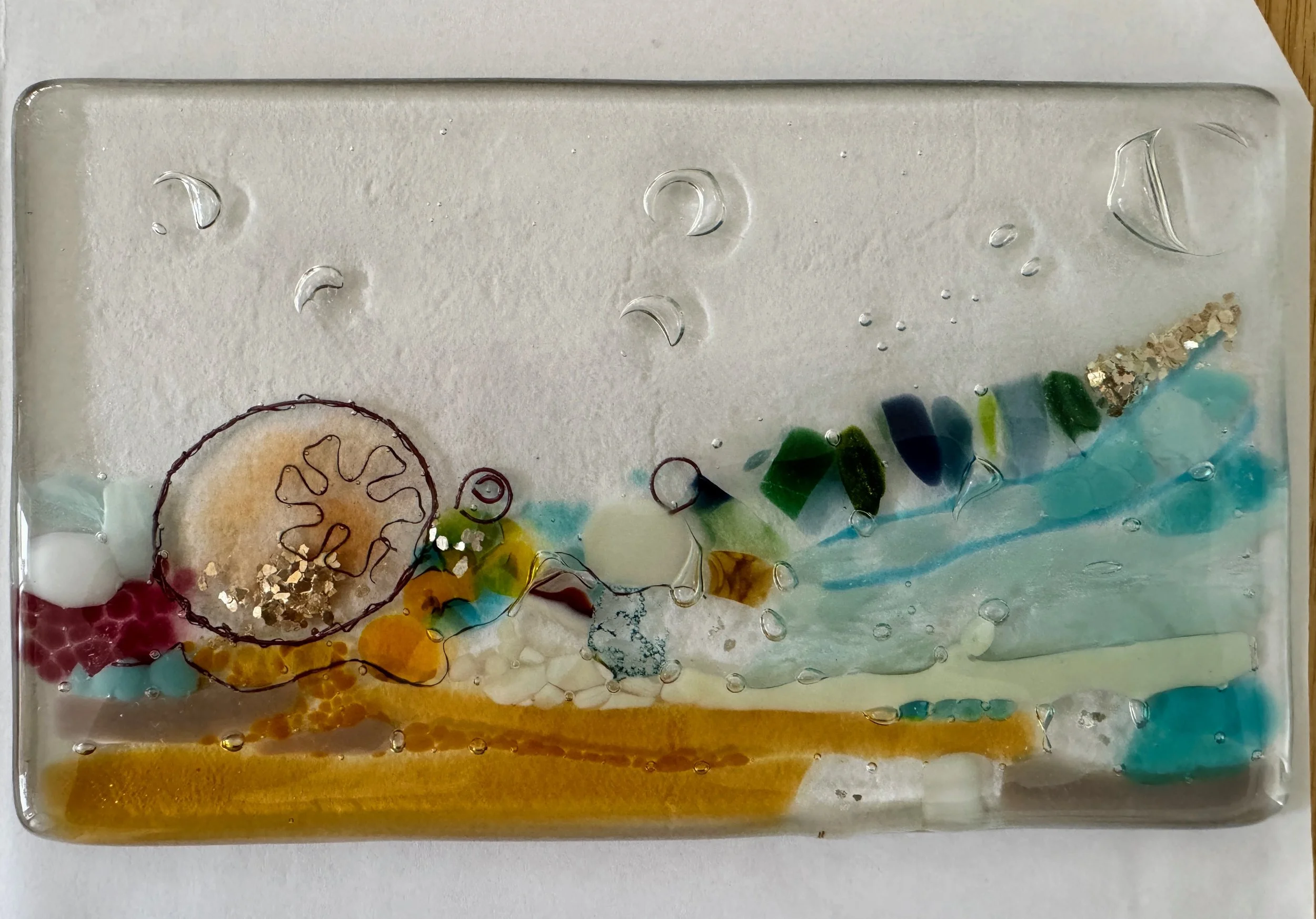 Artistic glass piece with colorful patterns and textures, including circular and linear designs in blue, green, yellow, and red on a transparent background.