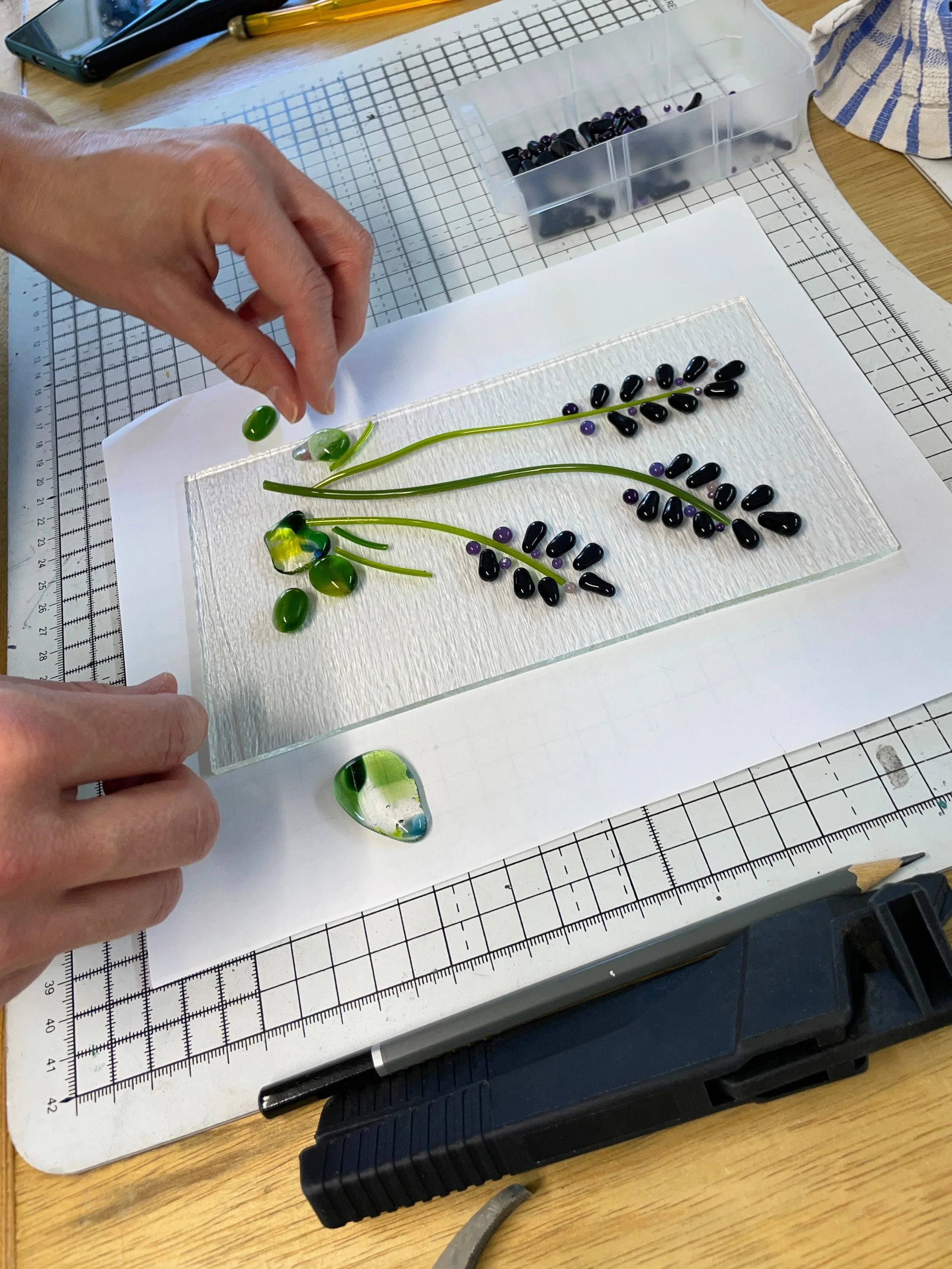 Person arranging glass pieces into a floral pattern on a cutting mat with a design of lavender flowers.