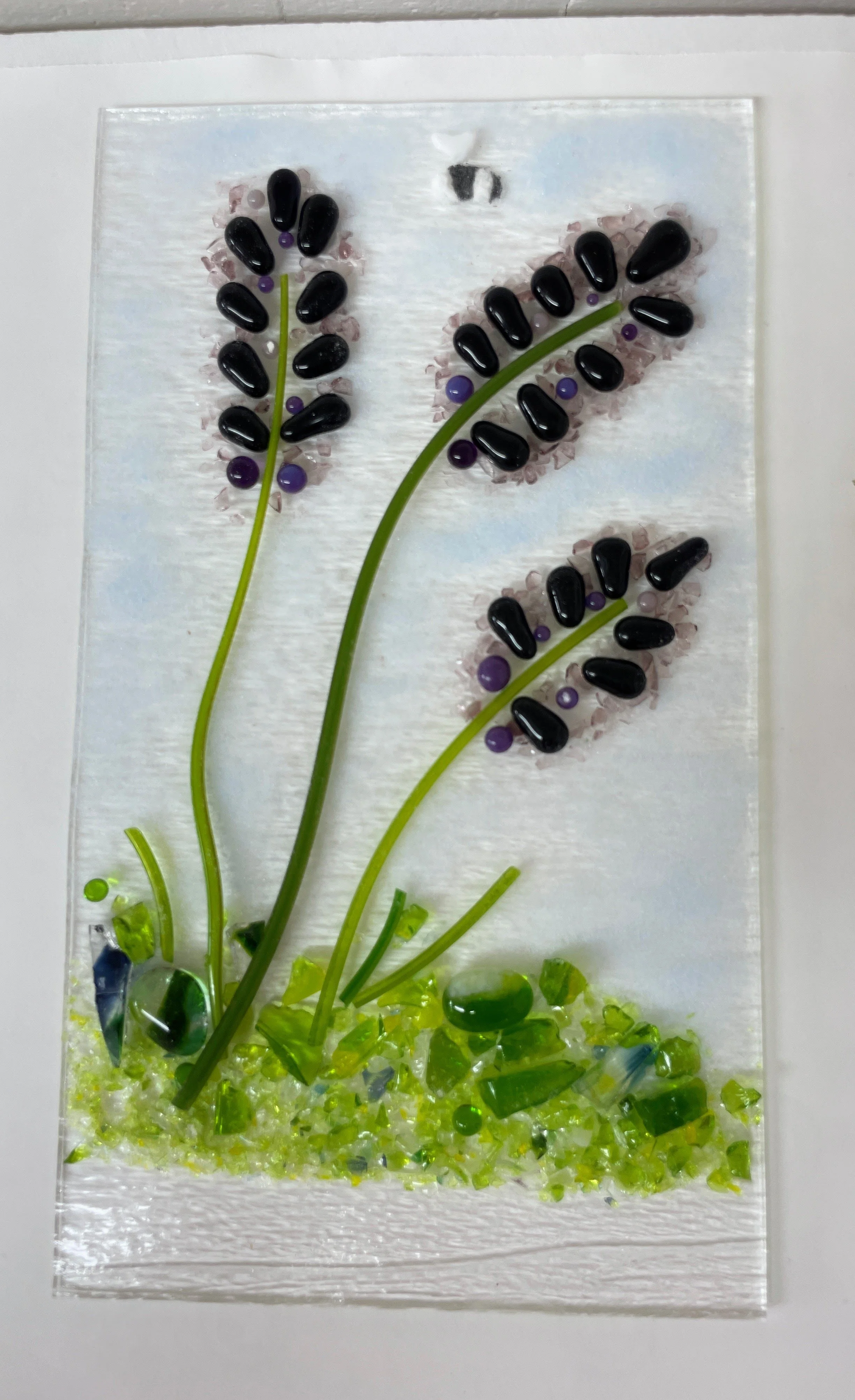 Fused glass art featuring three stylized flowers with black petals and purple accents on green stems, set against a white background and green glass shards at the base.