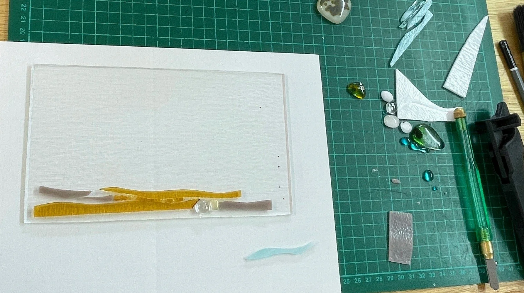 Glass artwork in progress on a cutting mat with glass pieces, cutting tools, and a white base.