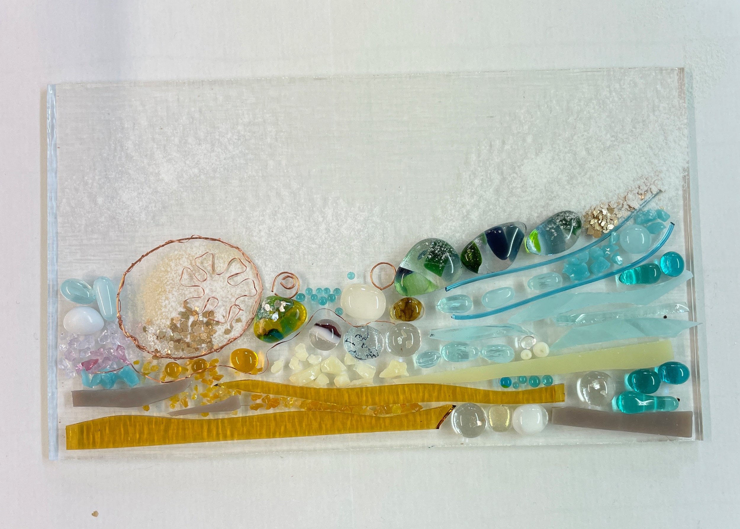 Abstract art on glass with colorful resin pieces, including blue, green, yellow, and brown elements, on a textured surface.