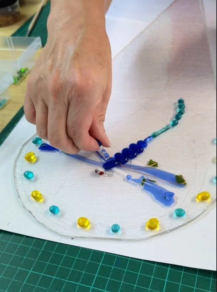 A person arranging colorful glass pieces on a circular template to create a mosaic design with blue glass elements resembling a dragonfly, surrounded by yellow and turquoise glass beads on a crafting surface.