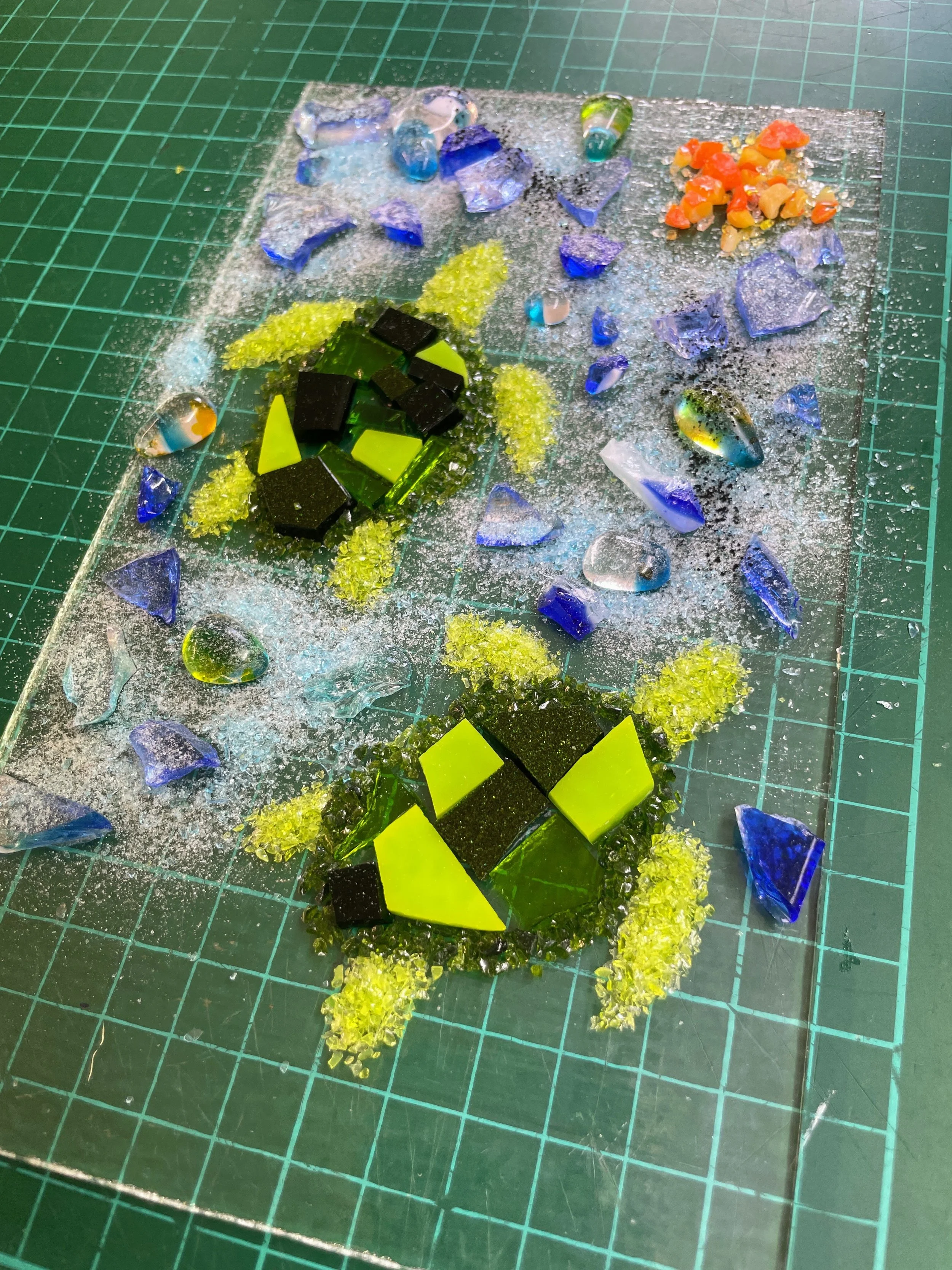 Colorful mosaic art depicting turtles with green and black tiles on a green grid cutting mat, surrounded by blue, orange, and clear glass pieces.
