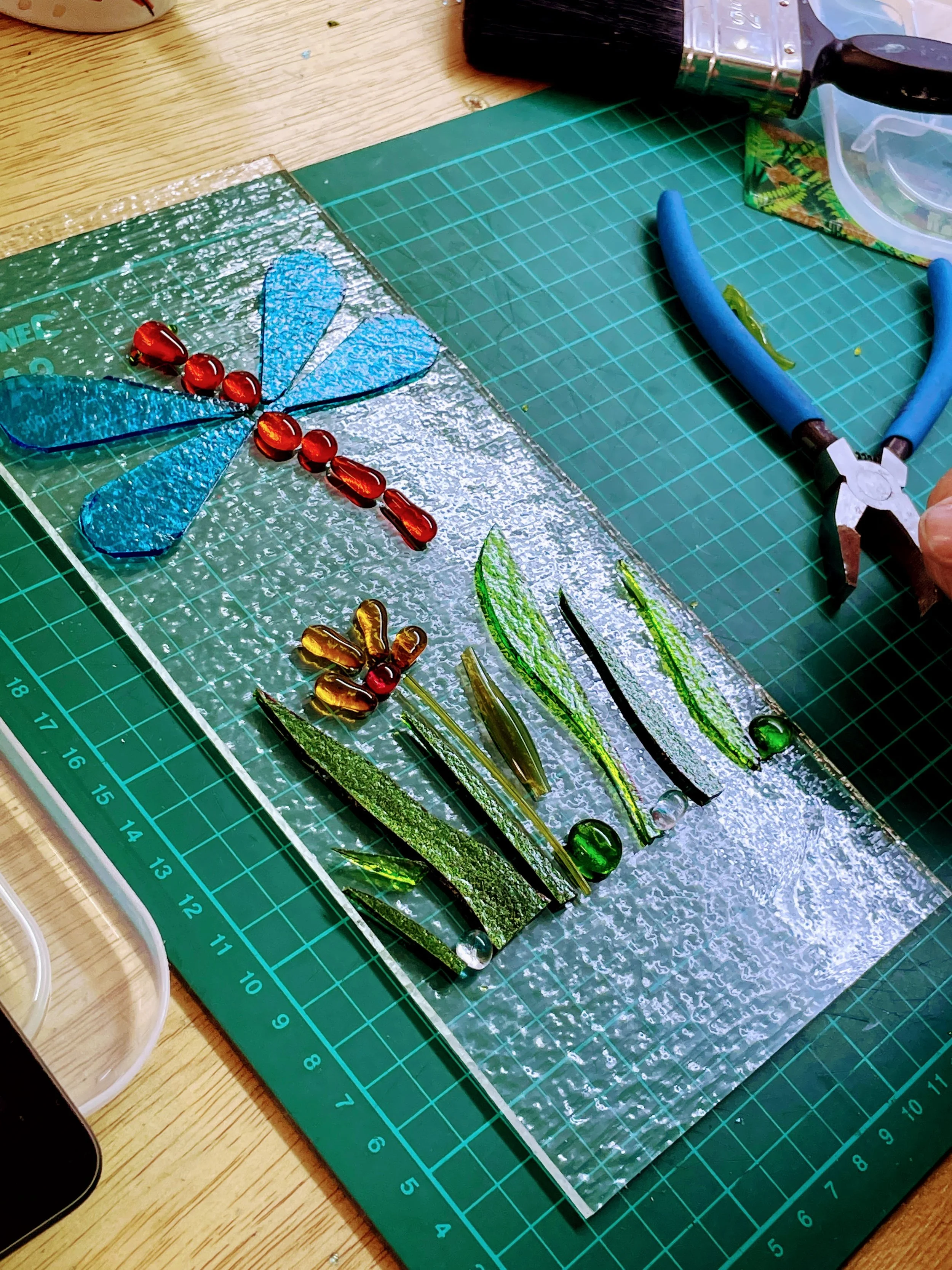 Stained glass artwork with blue dragonfly and green plants on cutting mat