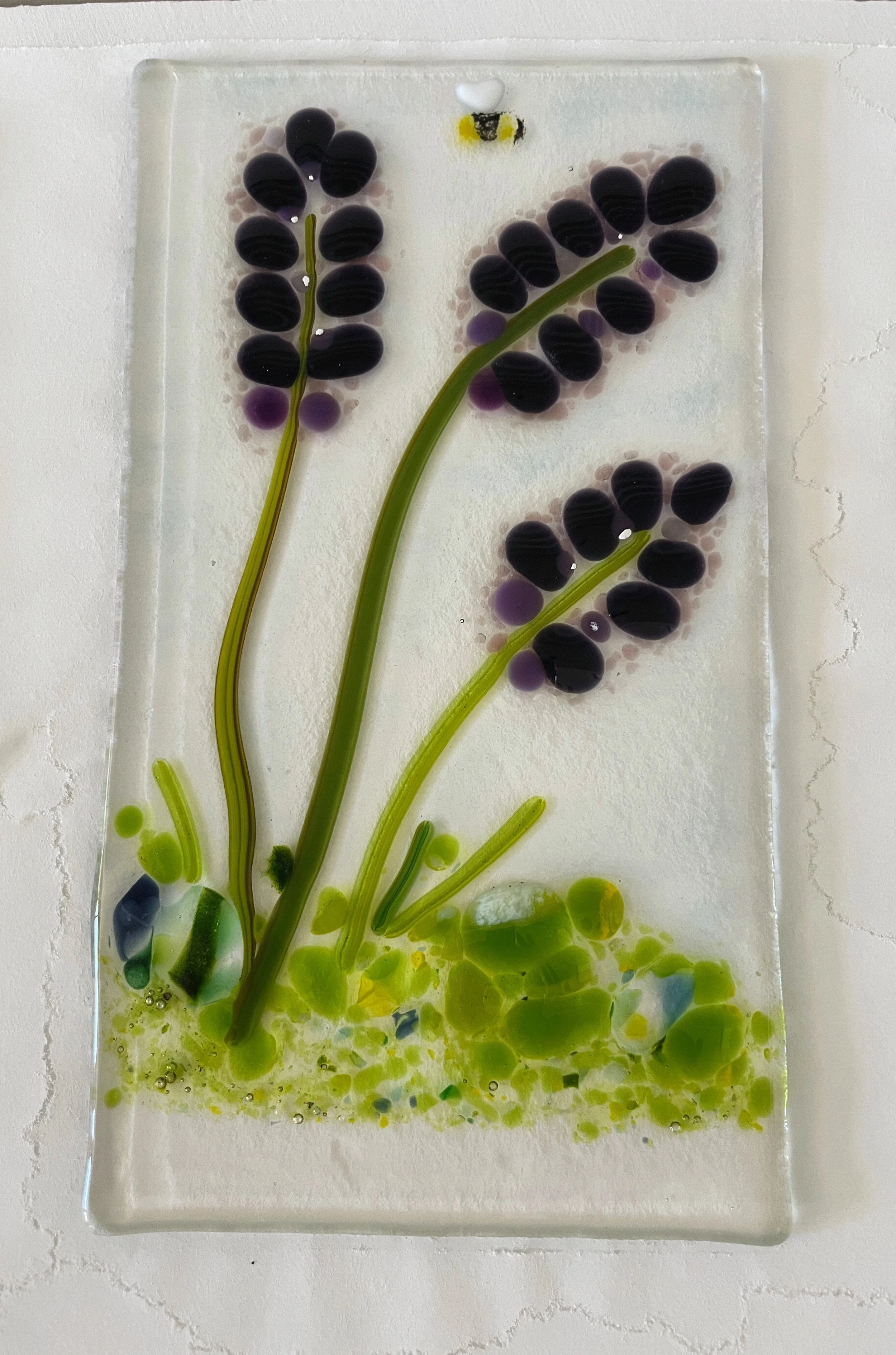 Handmade glass artwork depicting abstract purple flowers with green stems on a textured surface, featuring a small bee at the top.