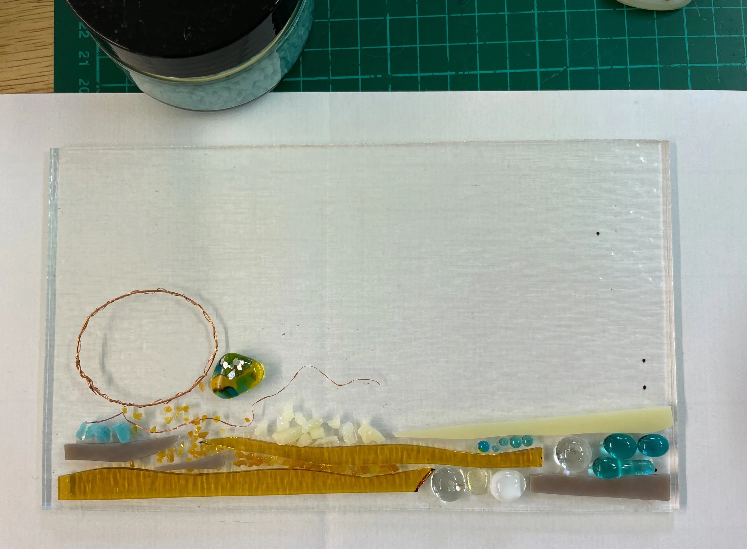 Abstract art piece on clear acrylic with various colored glass pieces, copper wire, and small decorative objects placed on a cutting mat background.