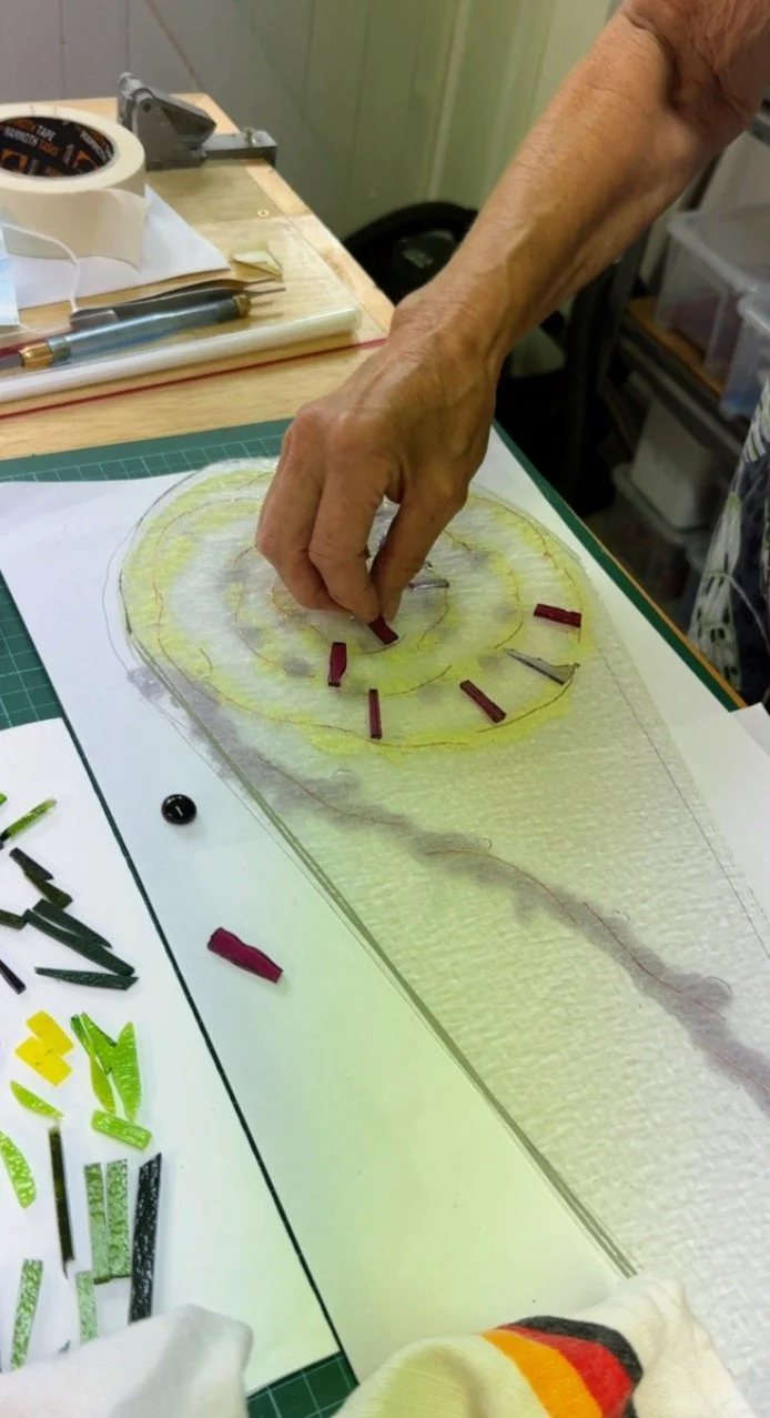 A person arranging small pieces of colored glass on a design template, possibly for a mosaic or stained glass project. Various tools and additional glass pieces are visible on the table.