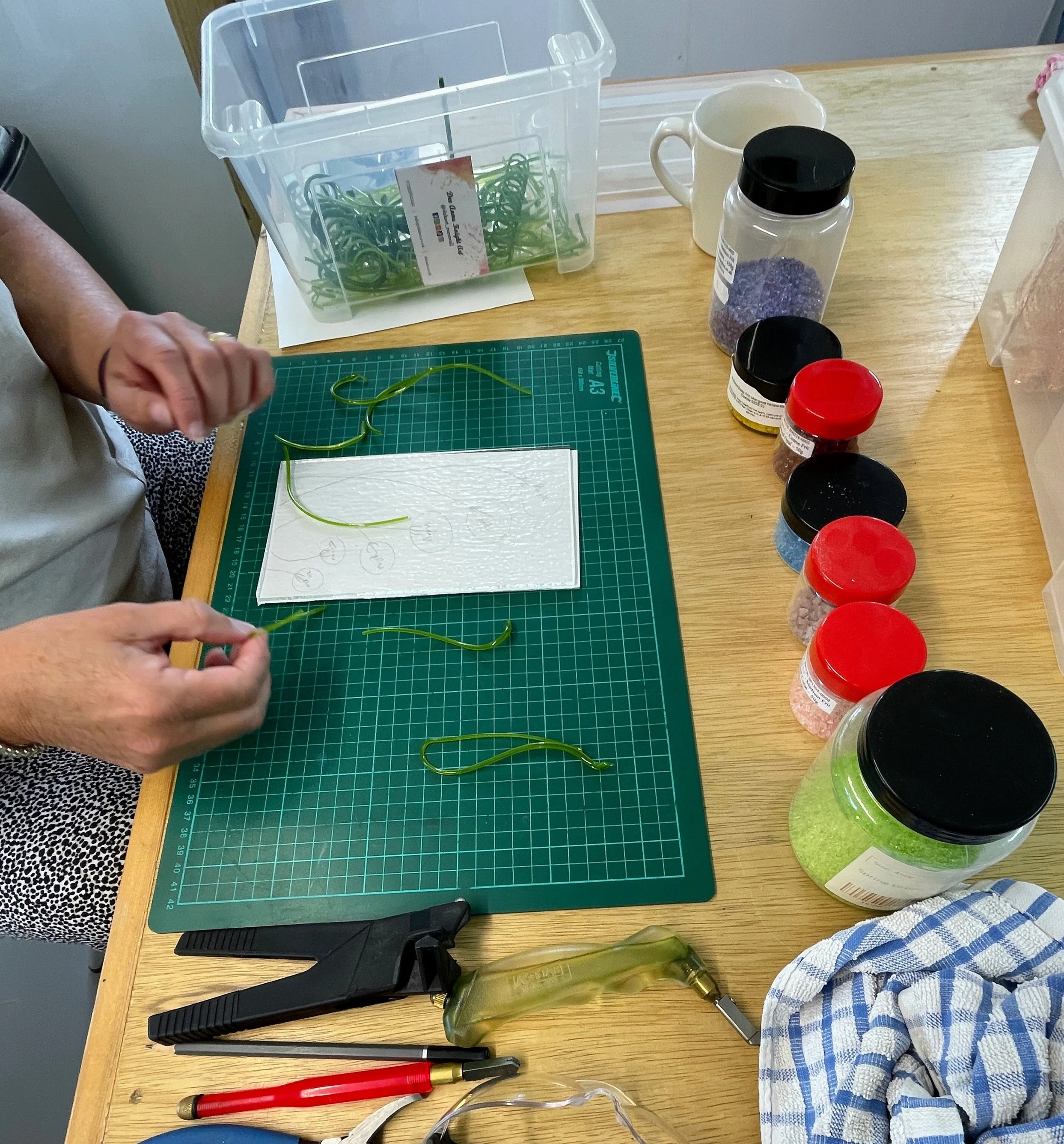A workspace with a person arranging green string-like material on a cutting mat. There are several containers with colored granules, tools, and a cloth on the wooden table.