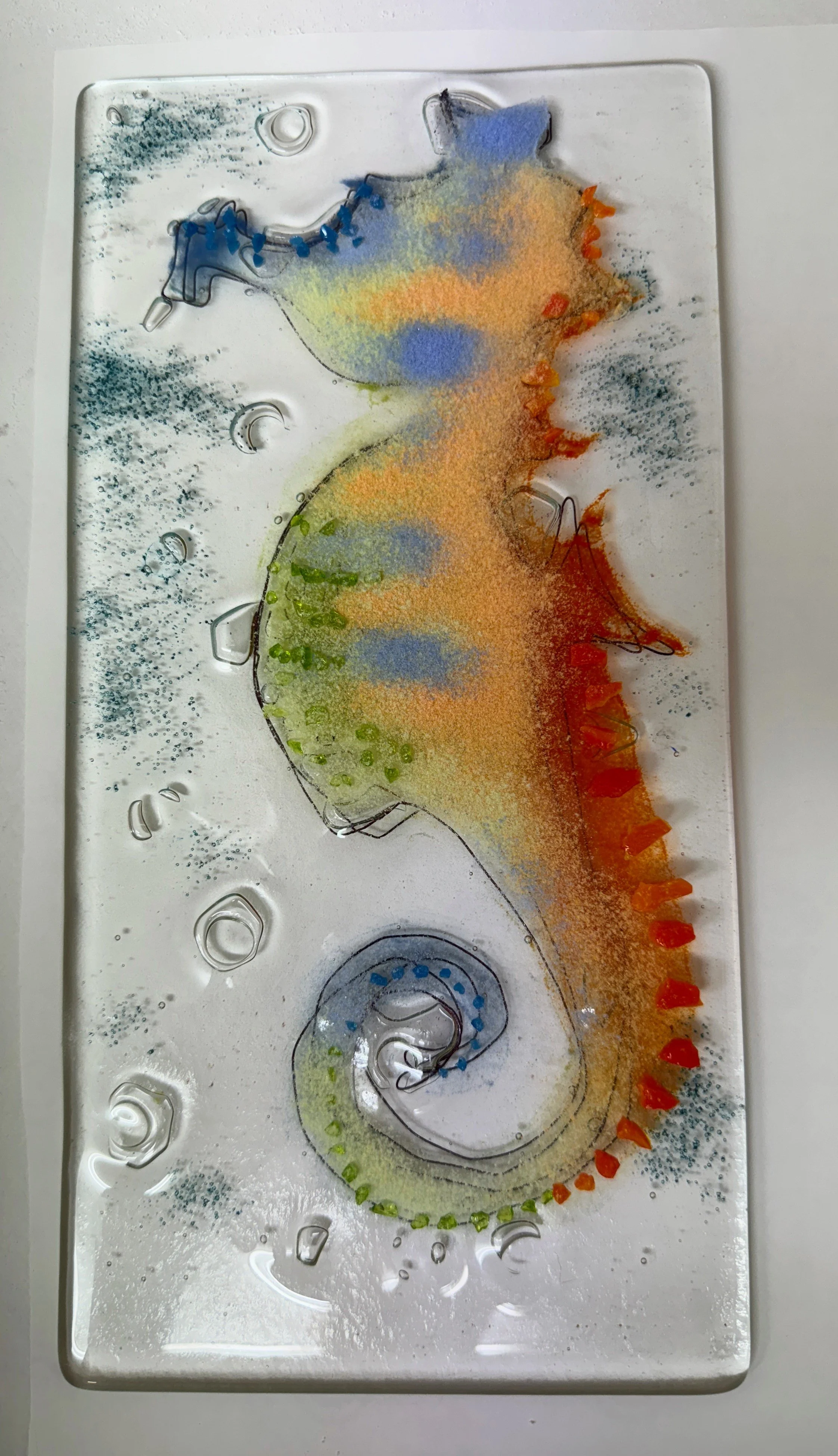 Colorful glass artwork featuring a seahorse with blue, orange, green, and yellow details on a clear background with textured elements.