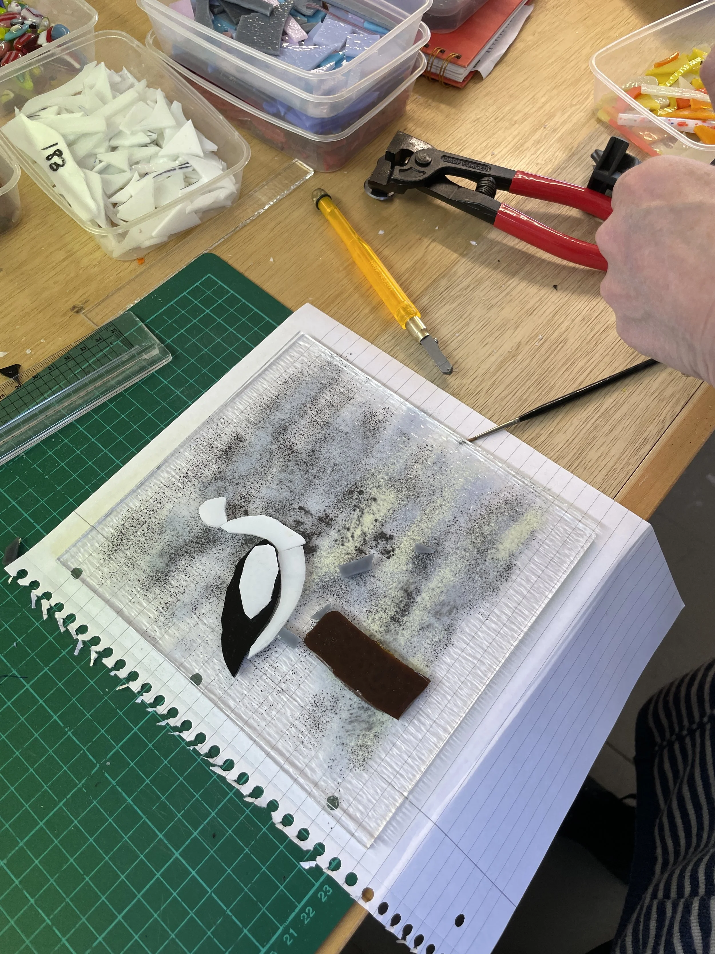 Crafting workspace with mosaic pieces in plastic containers, a cutting tool, ruler, and a glass art project on a notebook.