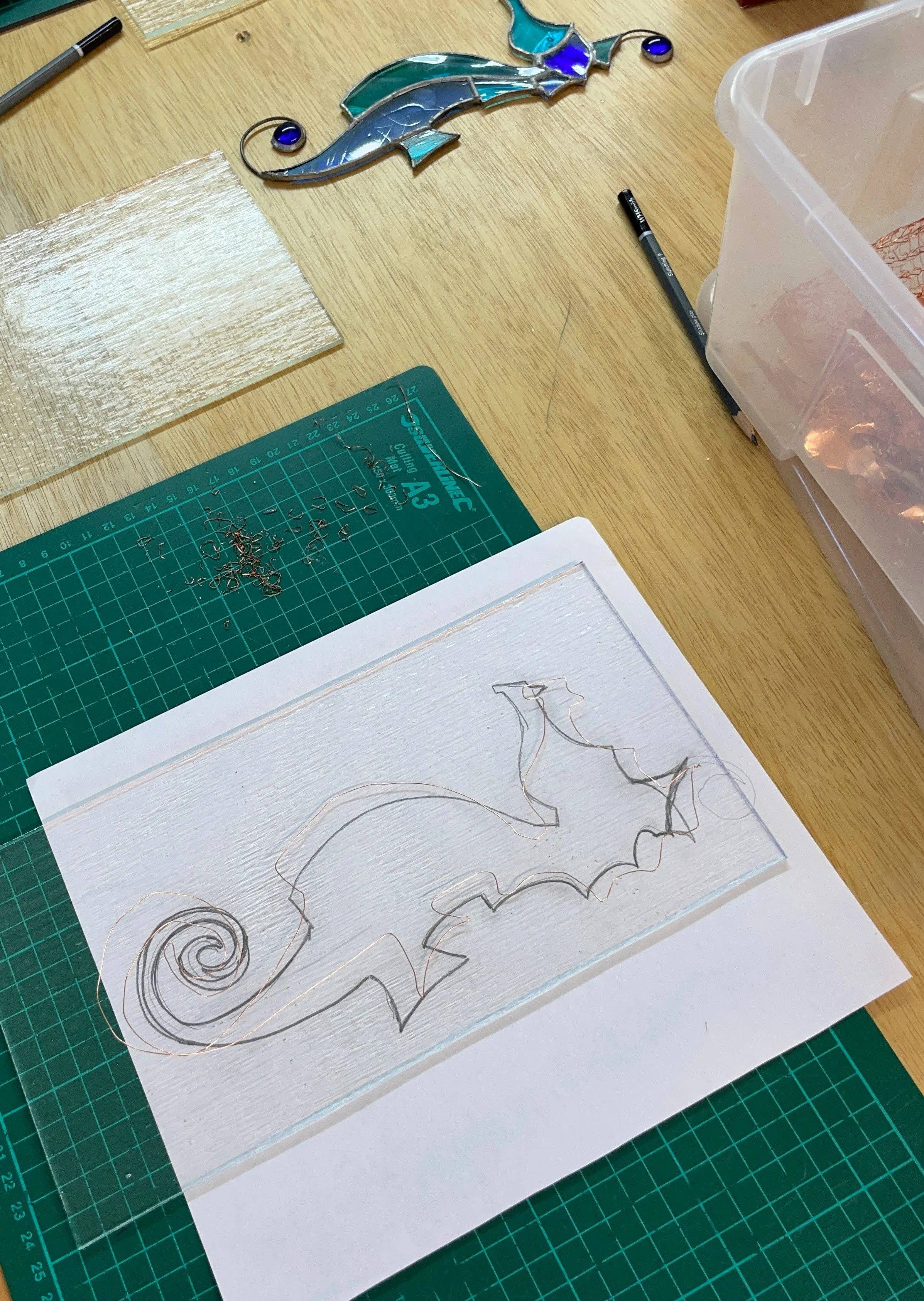 Glass art workbench with a sketched design of a chameleon, a cutting mat with metal scraps, a completed dolphin mosaic, and tools.