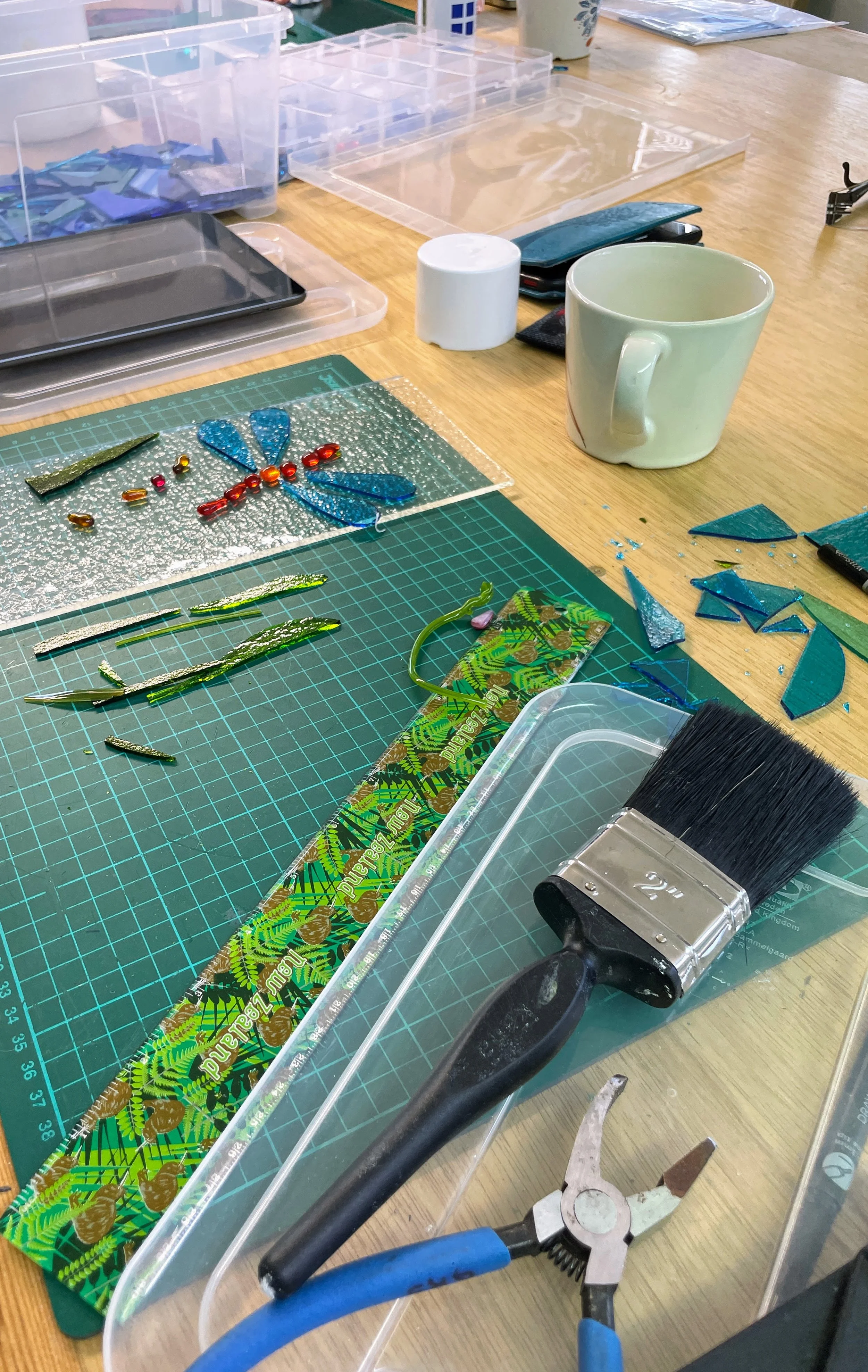 Crafting workspace with glass pieces, a green cutting mat, pliers, brush, ruler, and a mug. A dragonfly design made of colorful glass pieces is visible.