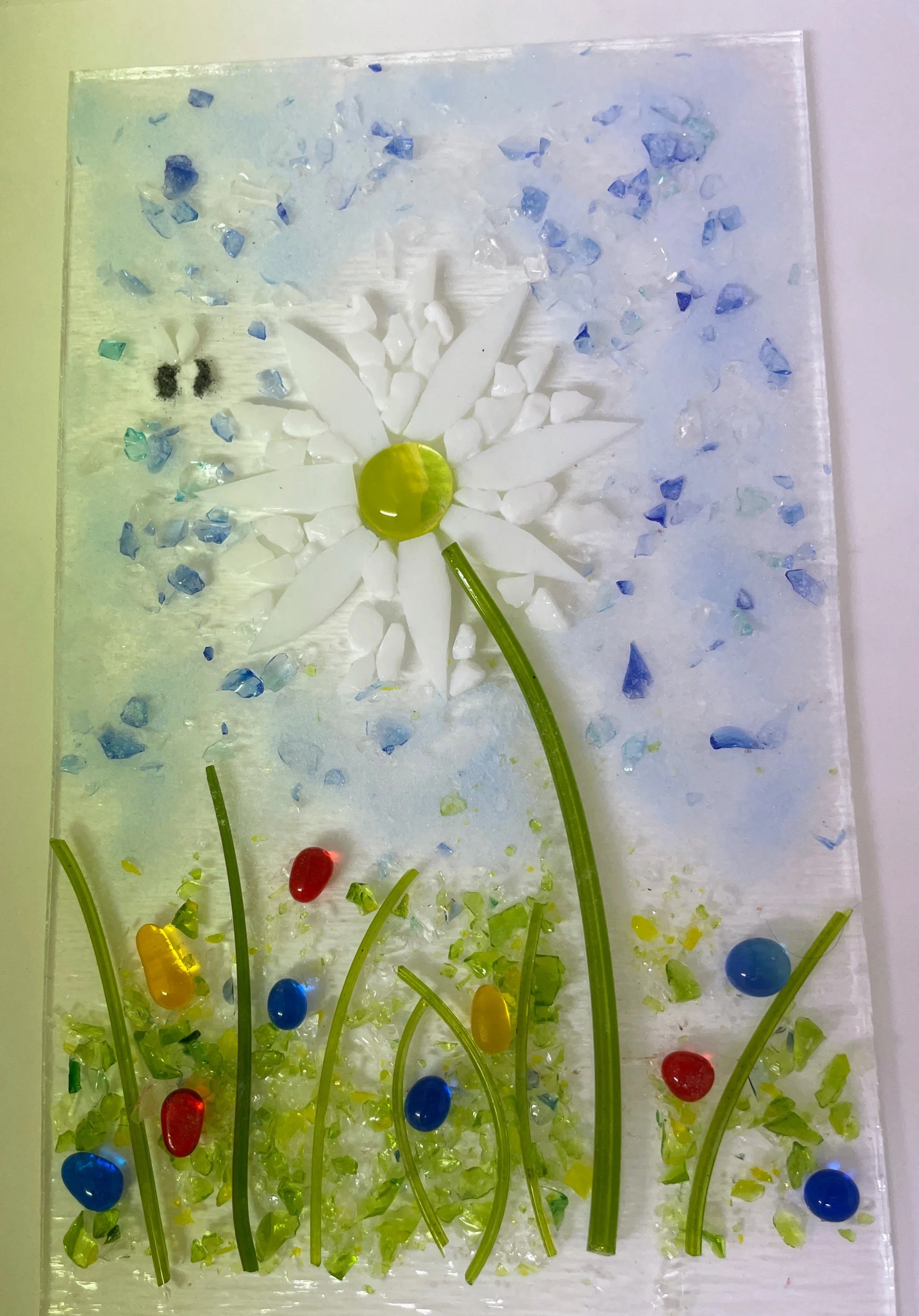 Mosaic art featuring a white daisy with a yellow center, green stems, and multi-colored stones representing grass and sky.