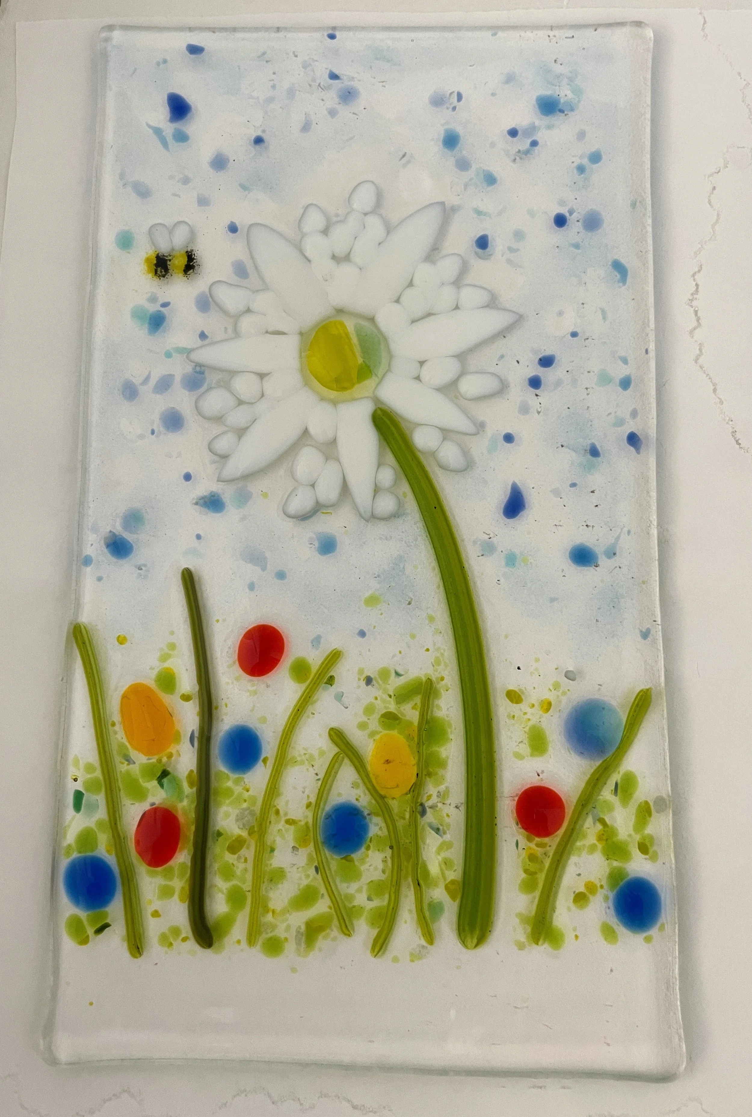 Decorative glass art panel featuring a white daisy with a yellow center, green stem, colorful dots resembling abstract flowers, and a small bee. Background has a textured, speckled pattern in blue hues.