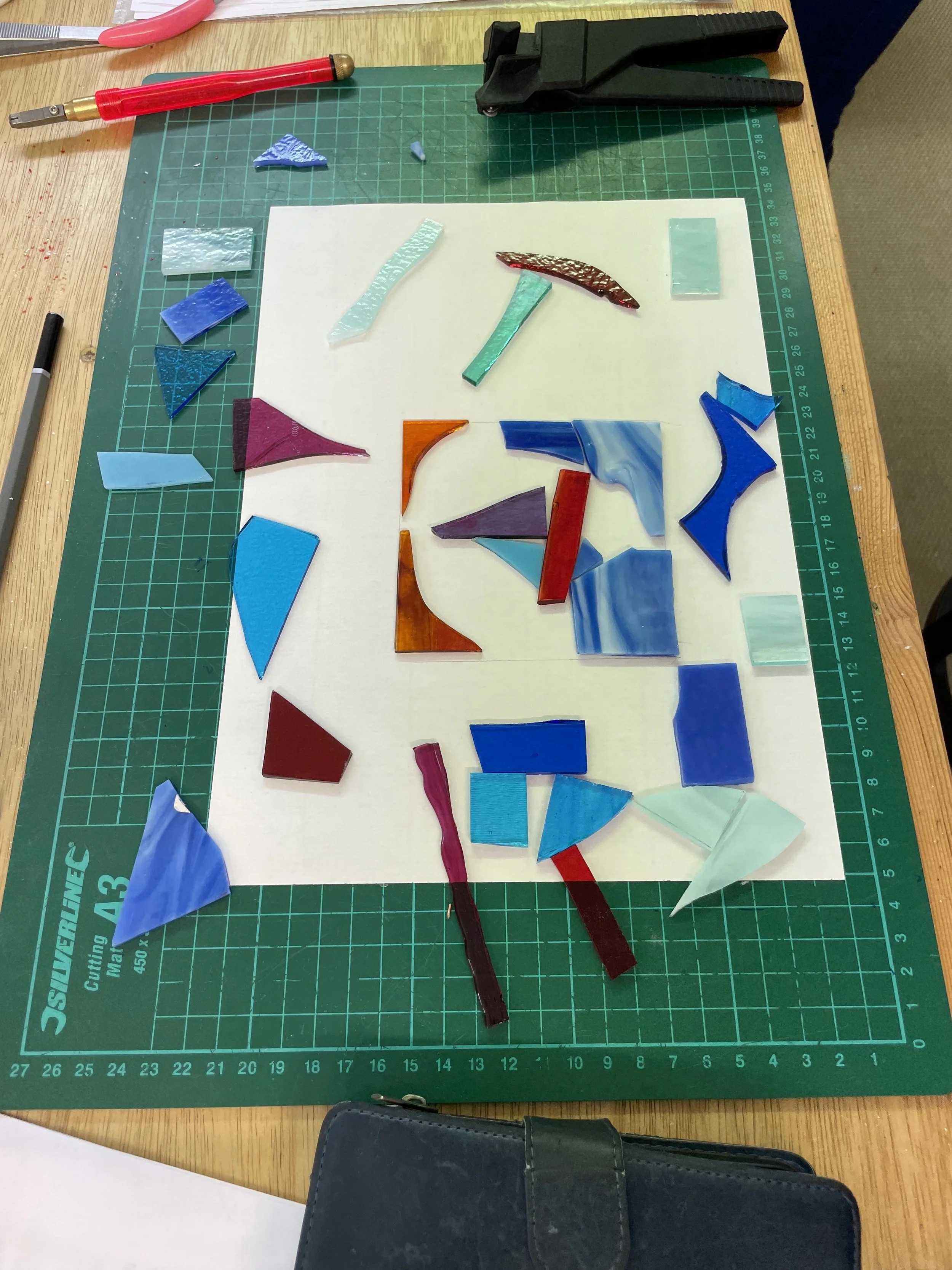 Colorful stained glass pieces on a green cutting mat with tools around, arranged on a white paper sheet.
