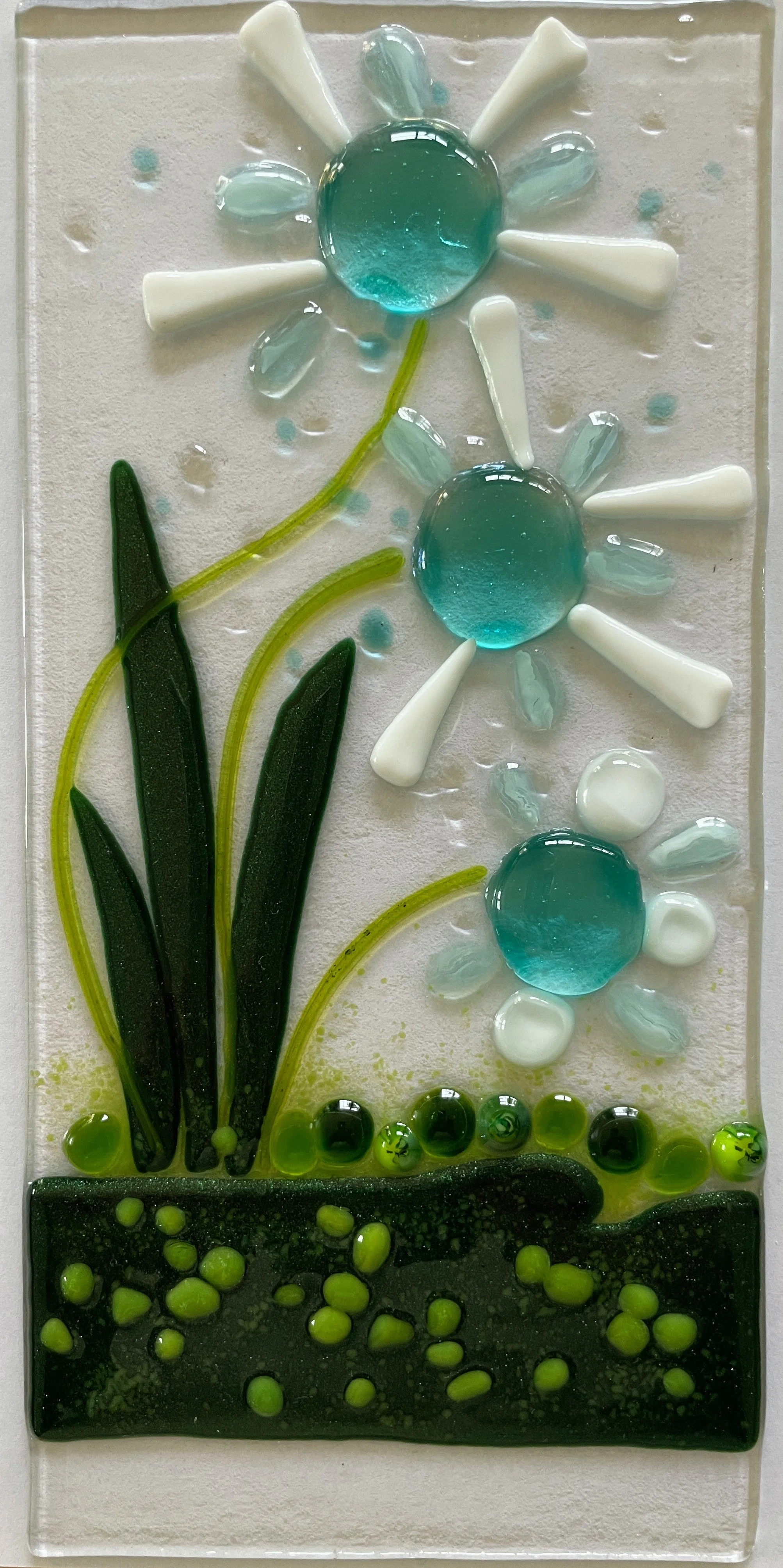 Glass art depicting flowers with blue centers and white petals, green stems and leaves on a textured background.