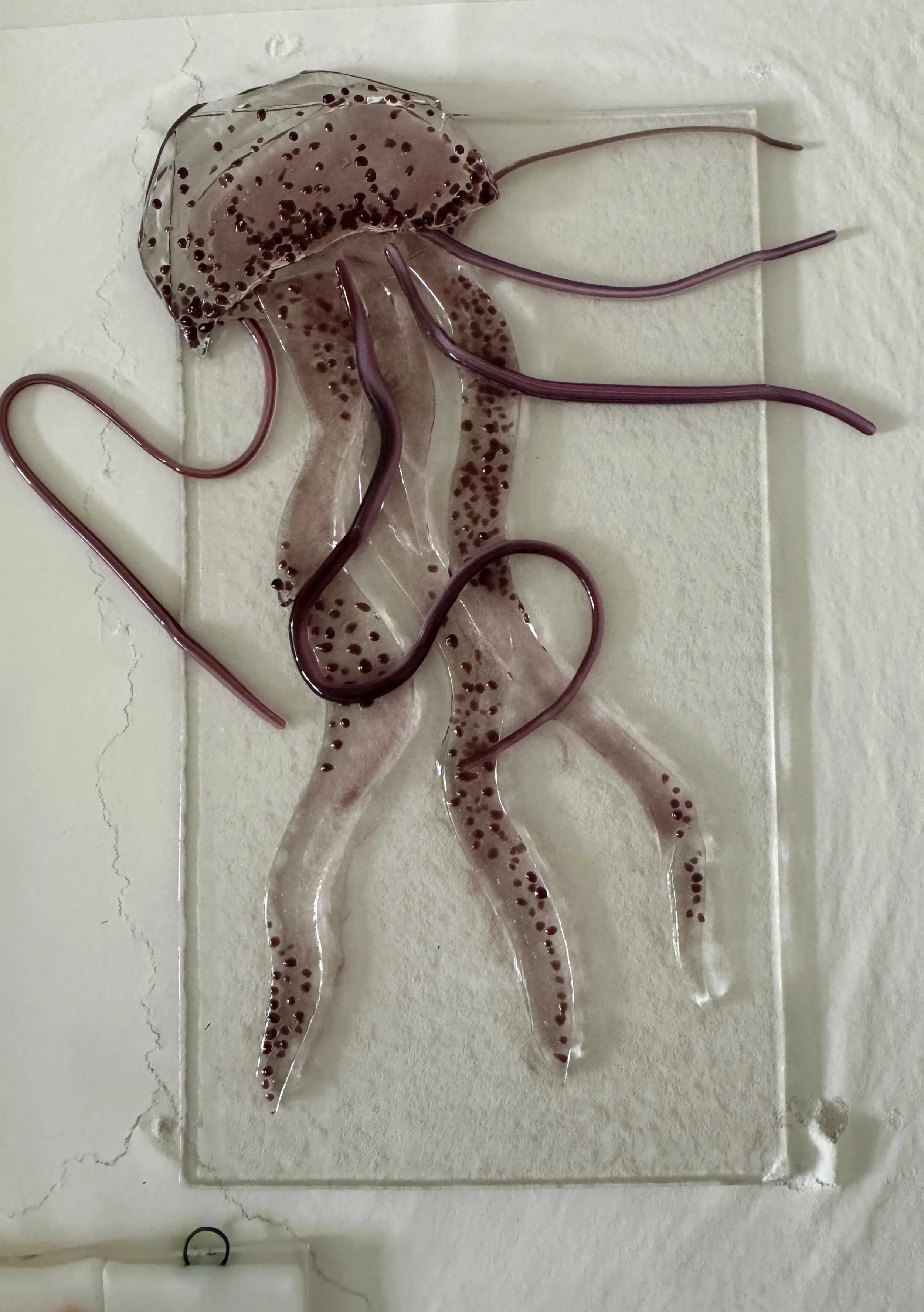 Glass art depiction of a jellyfish with purple tentacles and speckled body on a textured background.