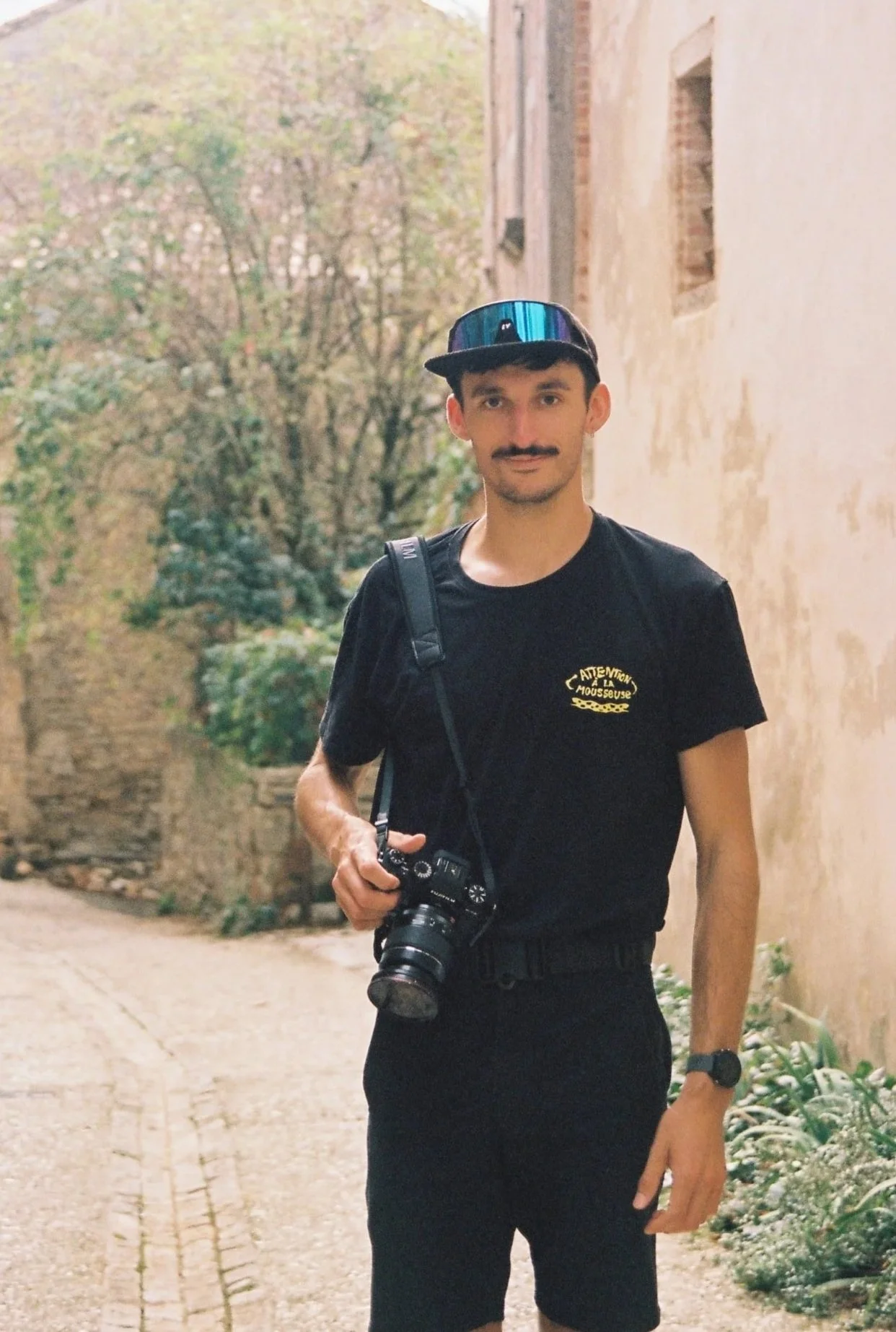 Un jeune homme avec un chapeau de casquette, moustache, portant un t-shirt noir avec un logo en français, porte un sac à dos et une caméra autour du cou. Gauthier Leclair
