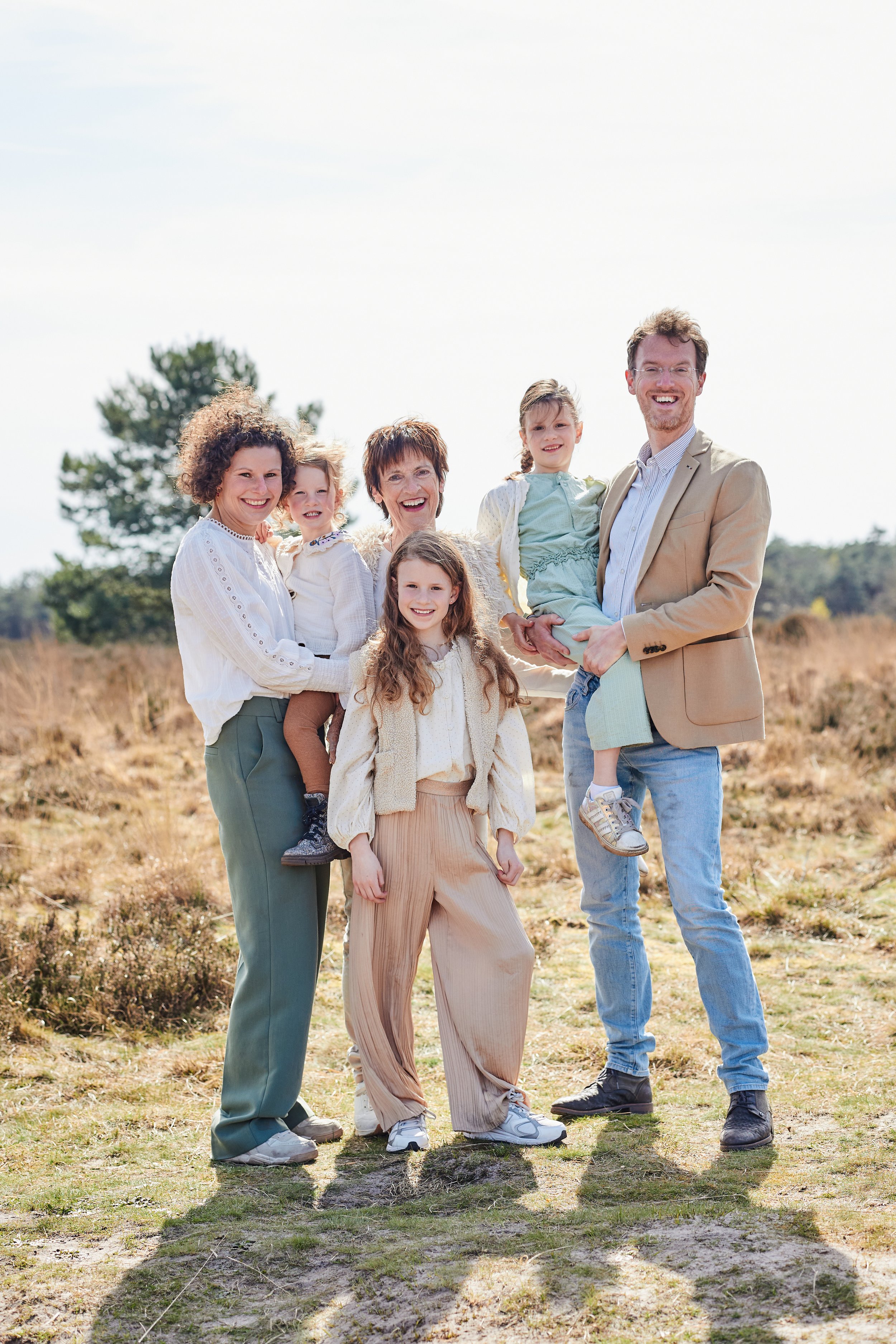Een familie van zeven mensen die buiten in een veld staan, glimlachend en poserend voor de camera. Ze lijken genieten van een zonnige dag.