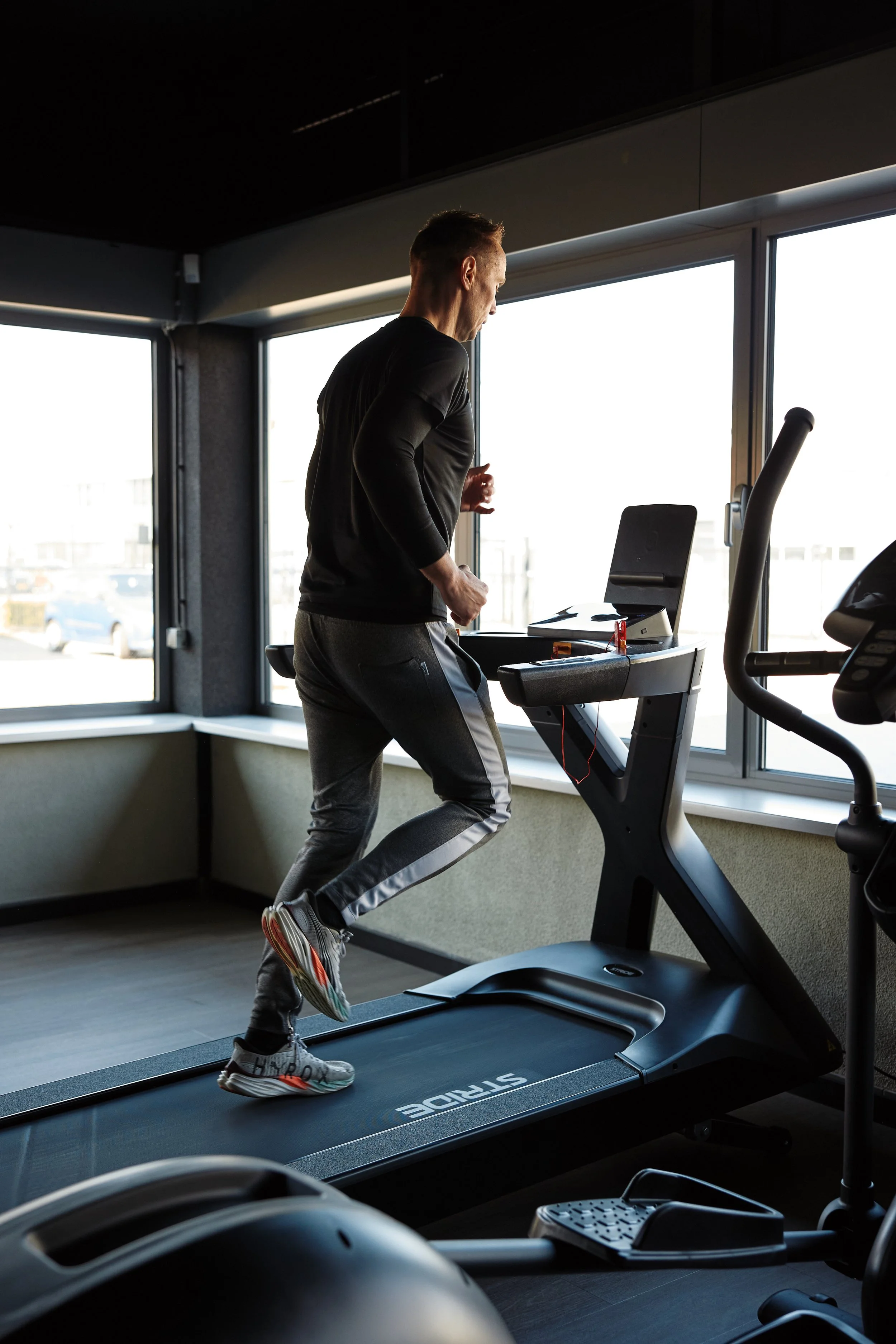 Man die hardloopt op een loopband in een fitnessruimte met grote ramen die uitzicht bieden op de buitenwereld.
