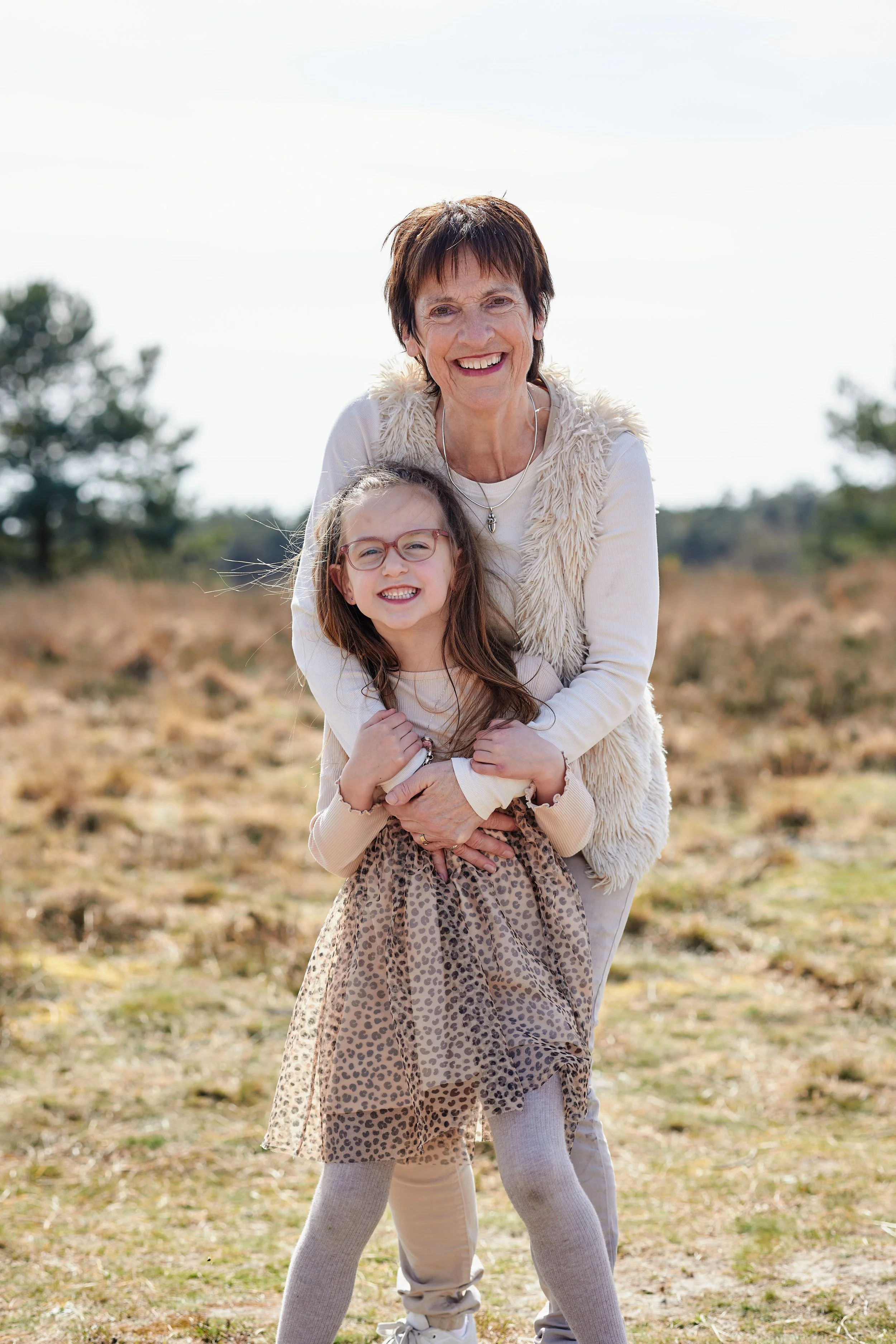 Een oudere vrouw en een jong meisje lachen samen buiten op een grasveld met bomen op de achtergrond, in een licht bewolkte hemel.