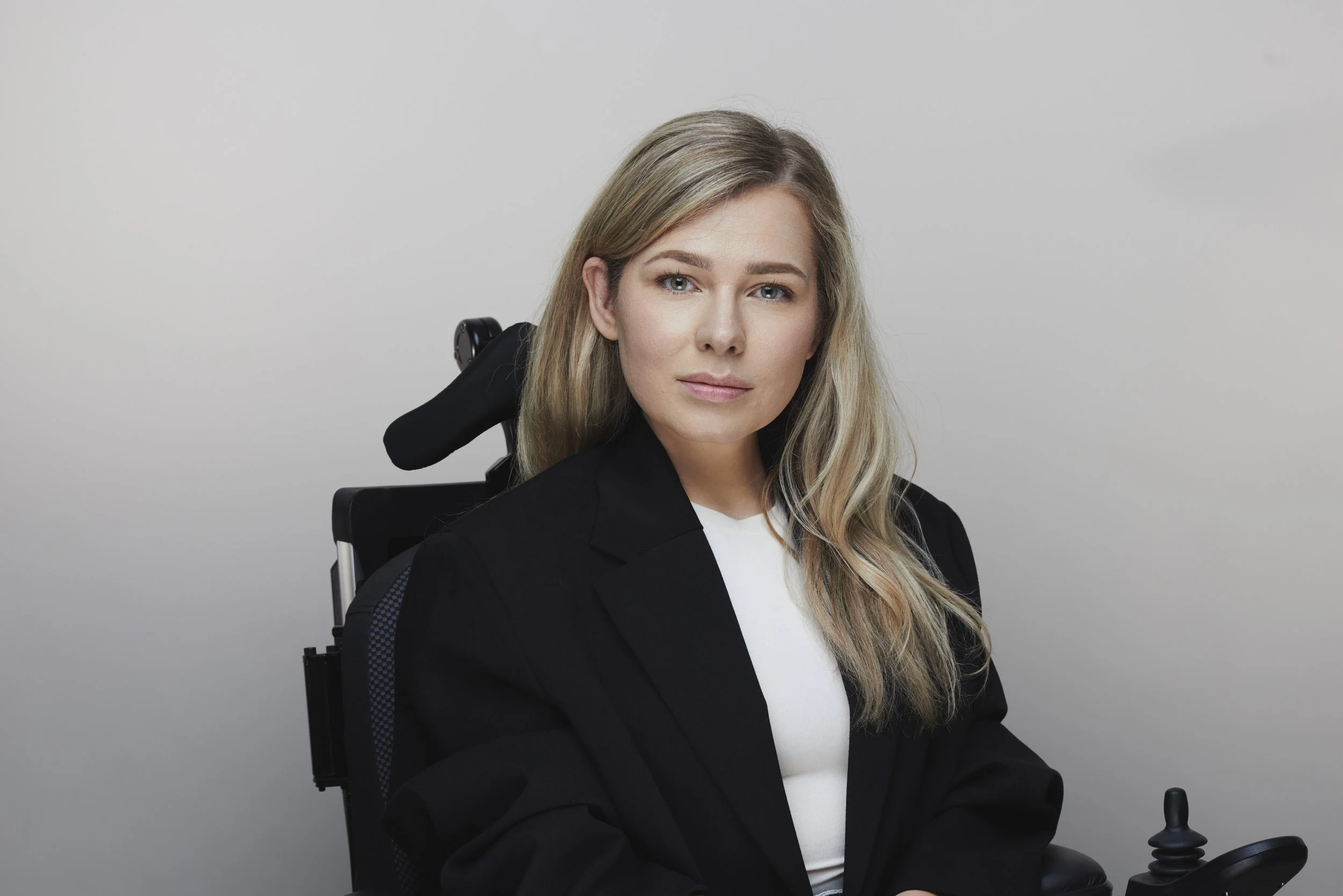 Een vrouw met lang blond haar zit in een rolstoel, draagt een zwarte blazer en een witte top, en kijkt in de camera. De achtergrond is grijs.