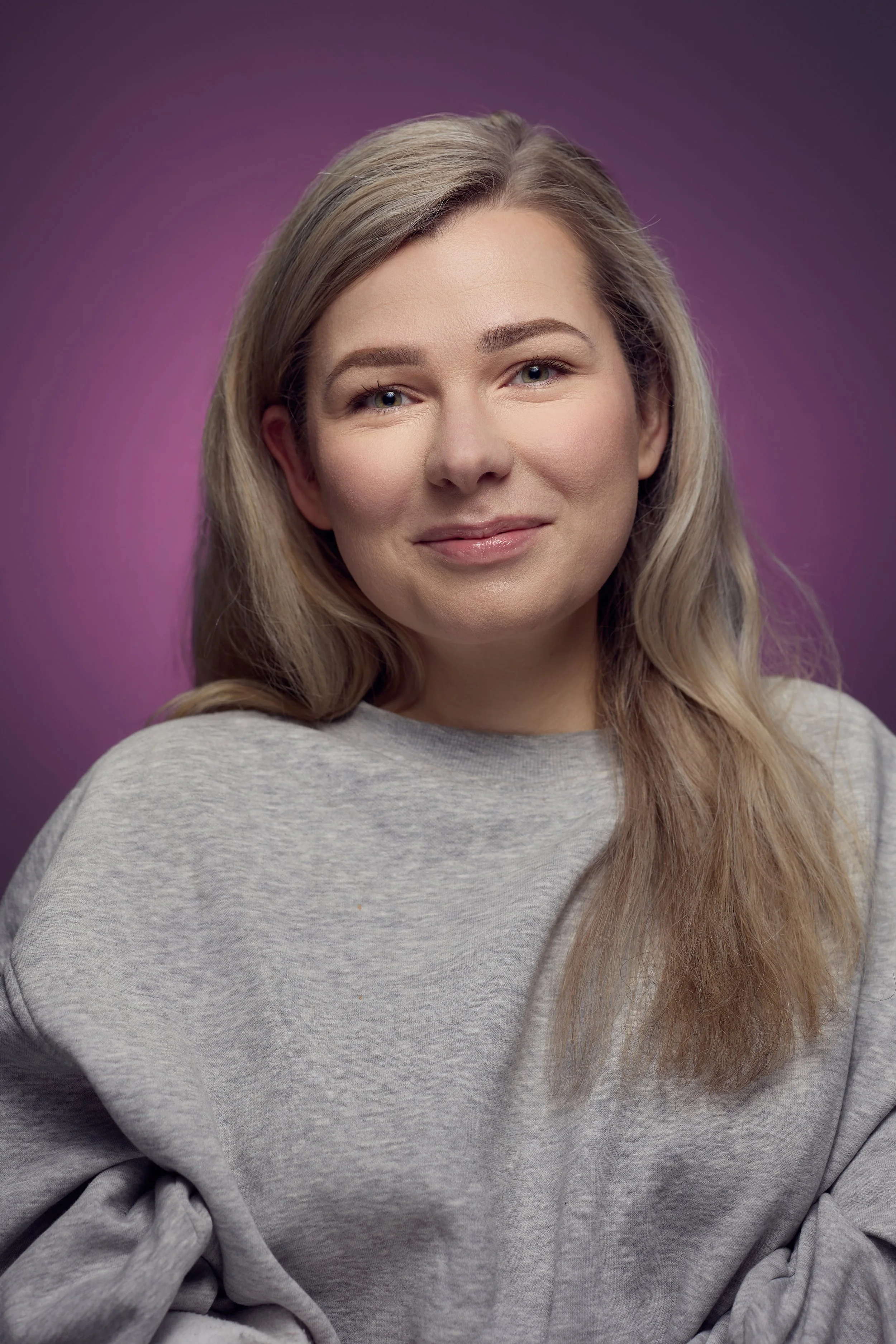 Close-up van een glimlachende vrouw met lang lichtbruin haar, gematteerde huid en blauwe ogen, gekleed in een grijs sweatshirt, tegen een paarse achtergrond.