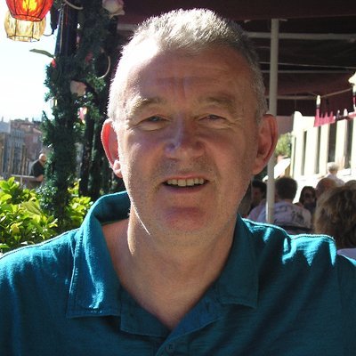 A smiling man with short gray hair wearing a blue shirt, sitting outdoors at a cafe or restaurant with plants and other people in the background.