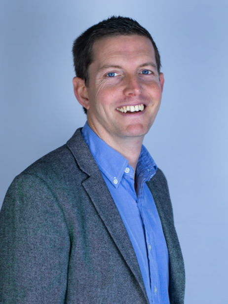 A smiling man with short dark hair and blue eyes, wearing a blue dress shirt and a gray blazer, standing against a light blue background.