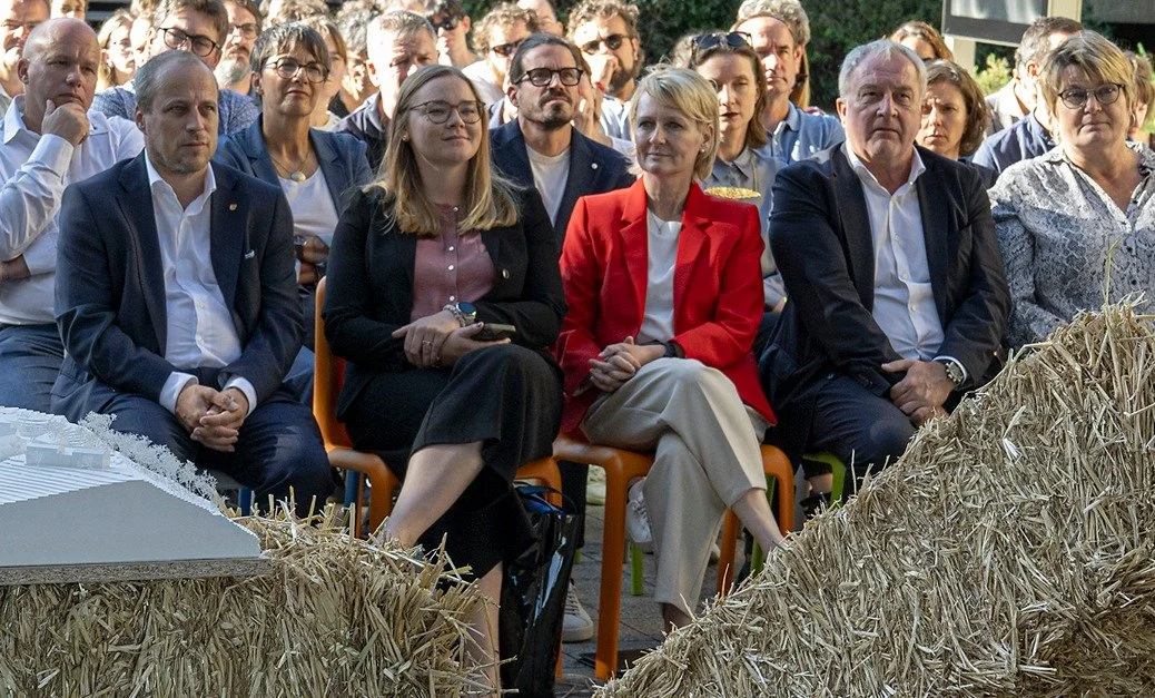 Groupe de personnes assises lors d'un événement extérieur, avec des végétaux en premier plan et des arbres en arrière-plan.