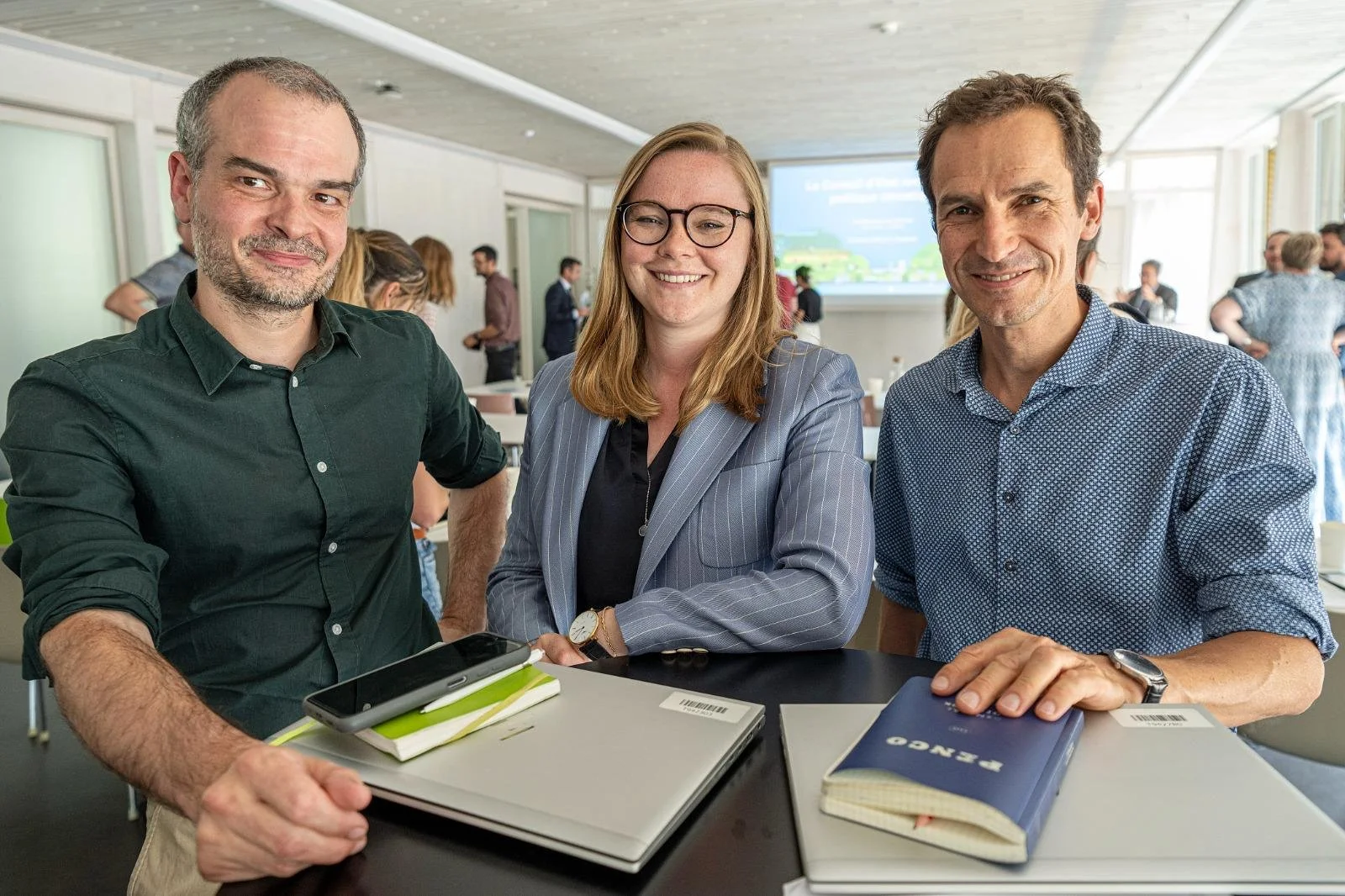 Trois personnes souriantes posant pour une photo lors d'une réunion ou d'un événement professionnel, avec d'autres personnes en arrière-plan